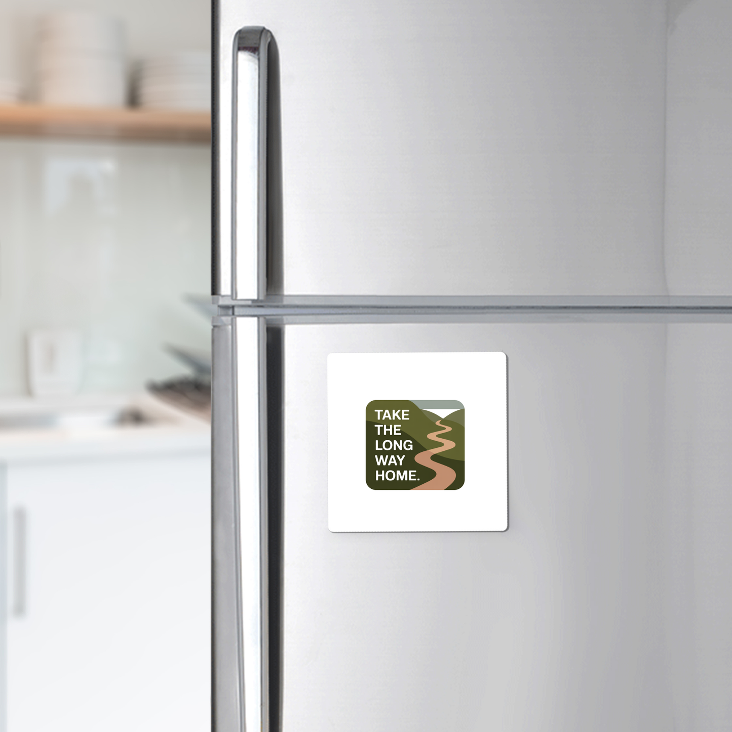 The Mirabilia Boutique "Take the Long Way Home Travel Magnet" is displayed on a stainless steel fridge, featuring a winding road illustration and text, set against nature-inspired décor with white dishes on shelves in a blurred kitchen.