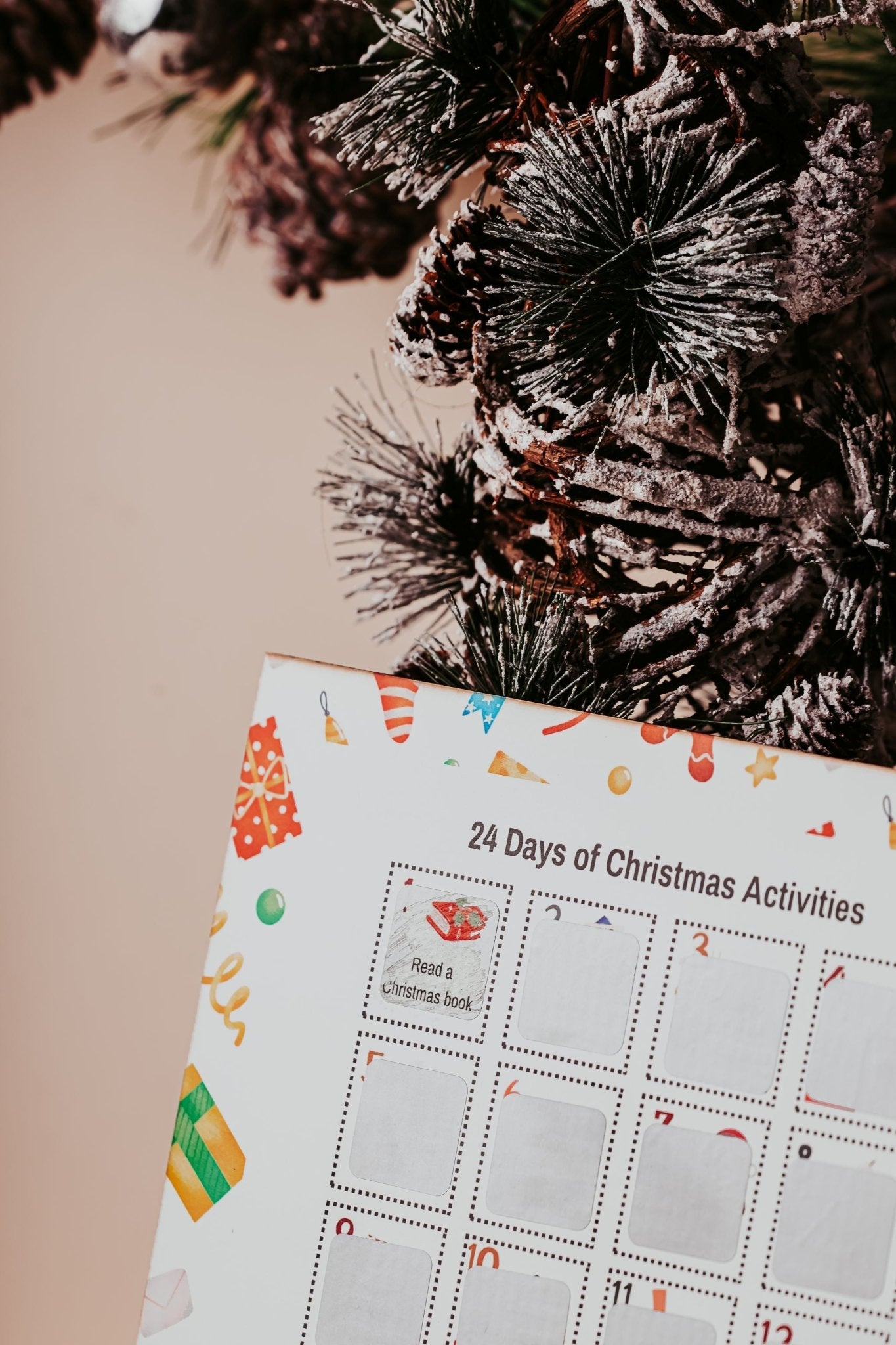 A snow-dusted pine branch sits beside the Mirabilia Boutique Scratch-Off Christmas Activity Calendar, a holiday countdown for kids featuring 25 festive family activities like “Read a Christmas book” and playful winter icons.