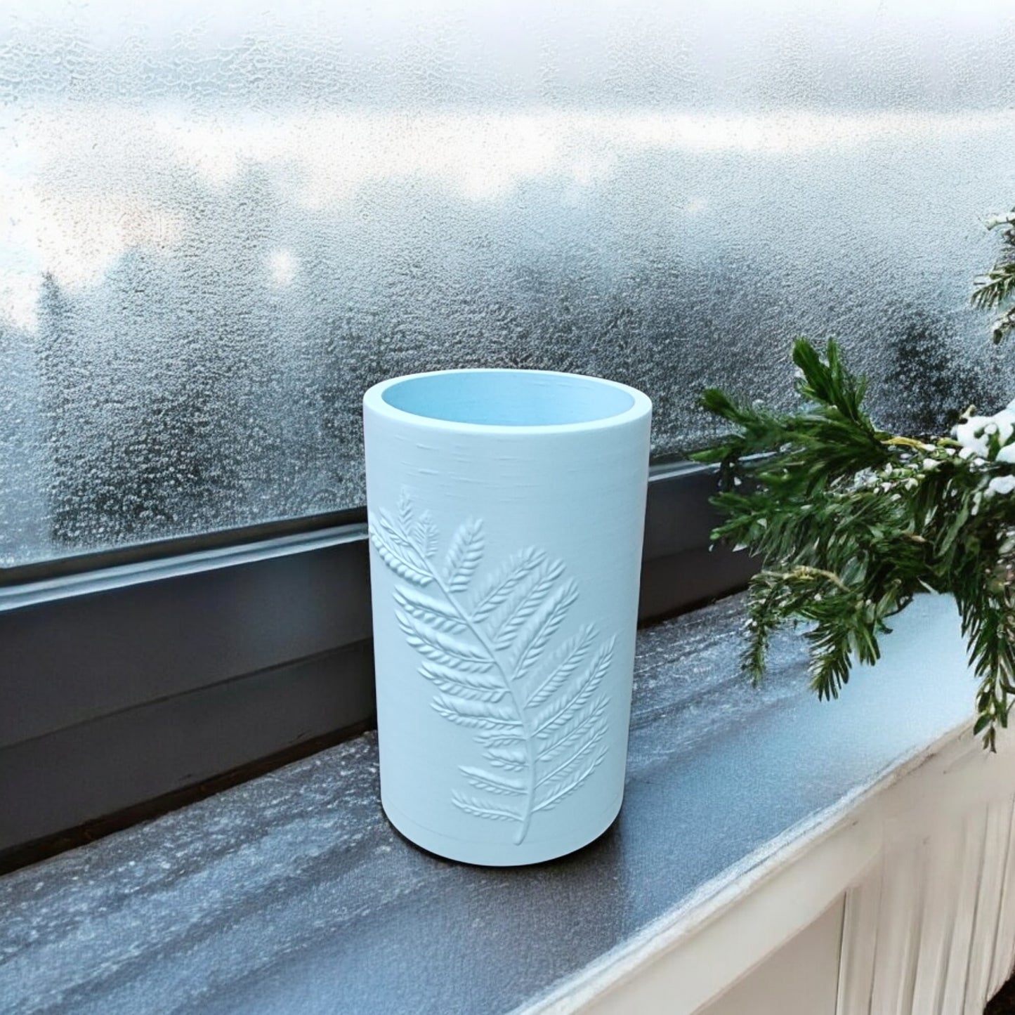 The Glacial Blue Winter Fern Vase – Woodland Accent by Mirabilia Boutique sits on a frosted windowsill, featuring a raised fern design and accompanied by a green pine branch visible on the right.