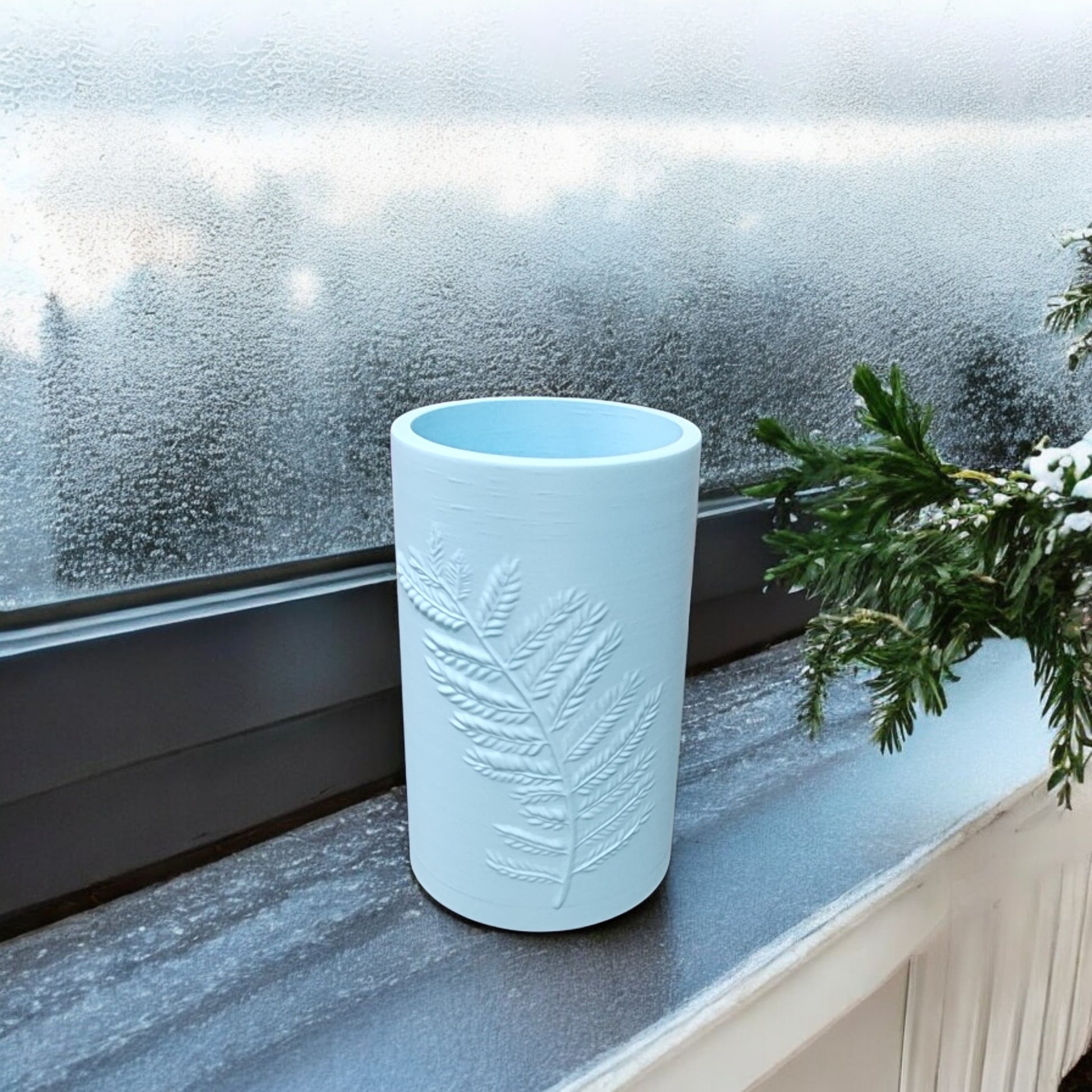 The Glacial Blue Winter Fern Vase – Woodland Accent by Mirabilia Boutique sits on a frosted windowsill, featuring a raised fern design and accompanied by a green pine branch visible on the right.