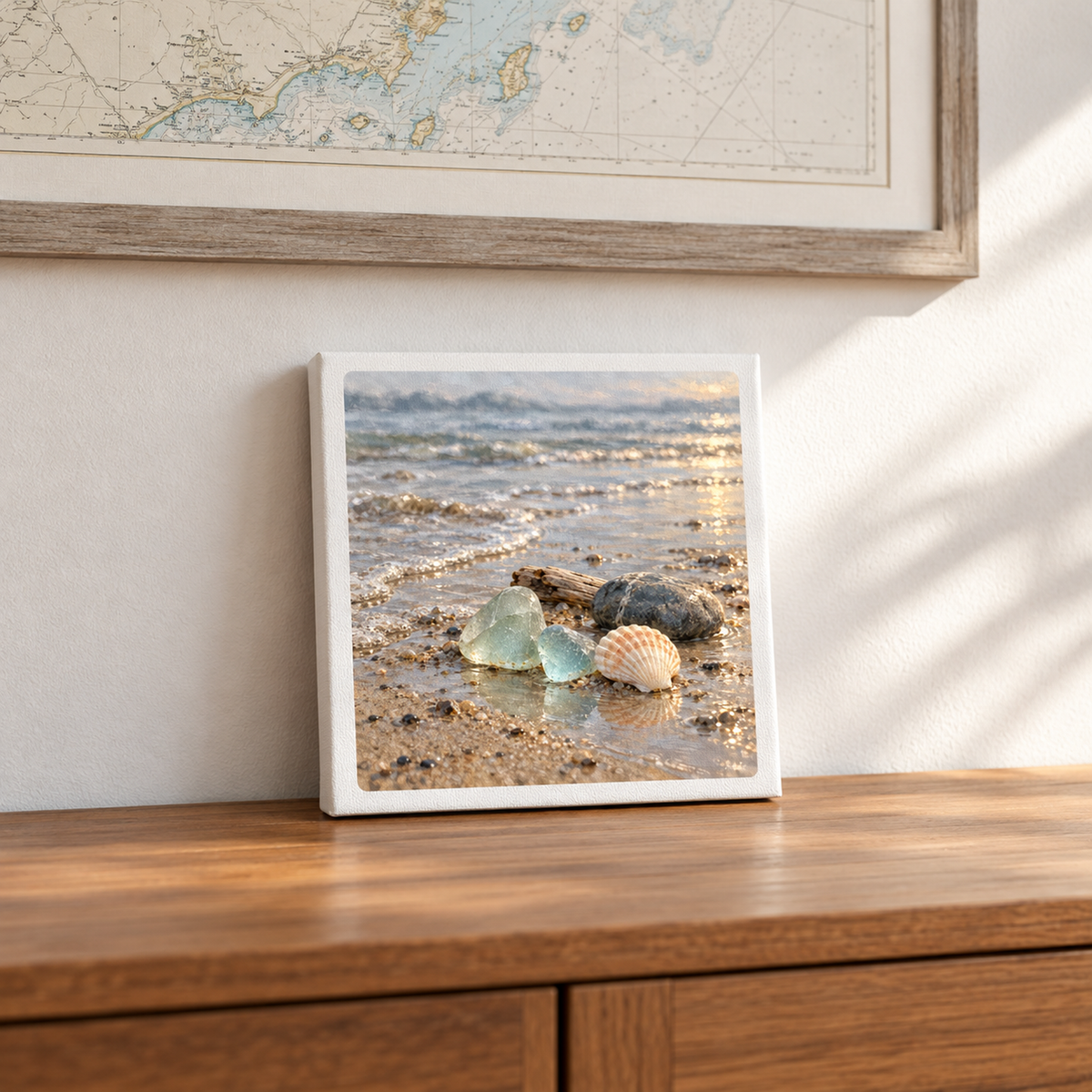 A Sea Glass Morning Canvas by Salt & Story rests on a wooden surface, showing a square print of sea glass, a seashell, and a stone on a wet, sunlit beach.