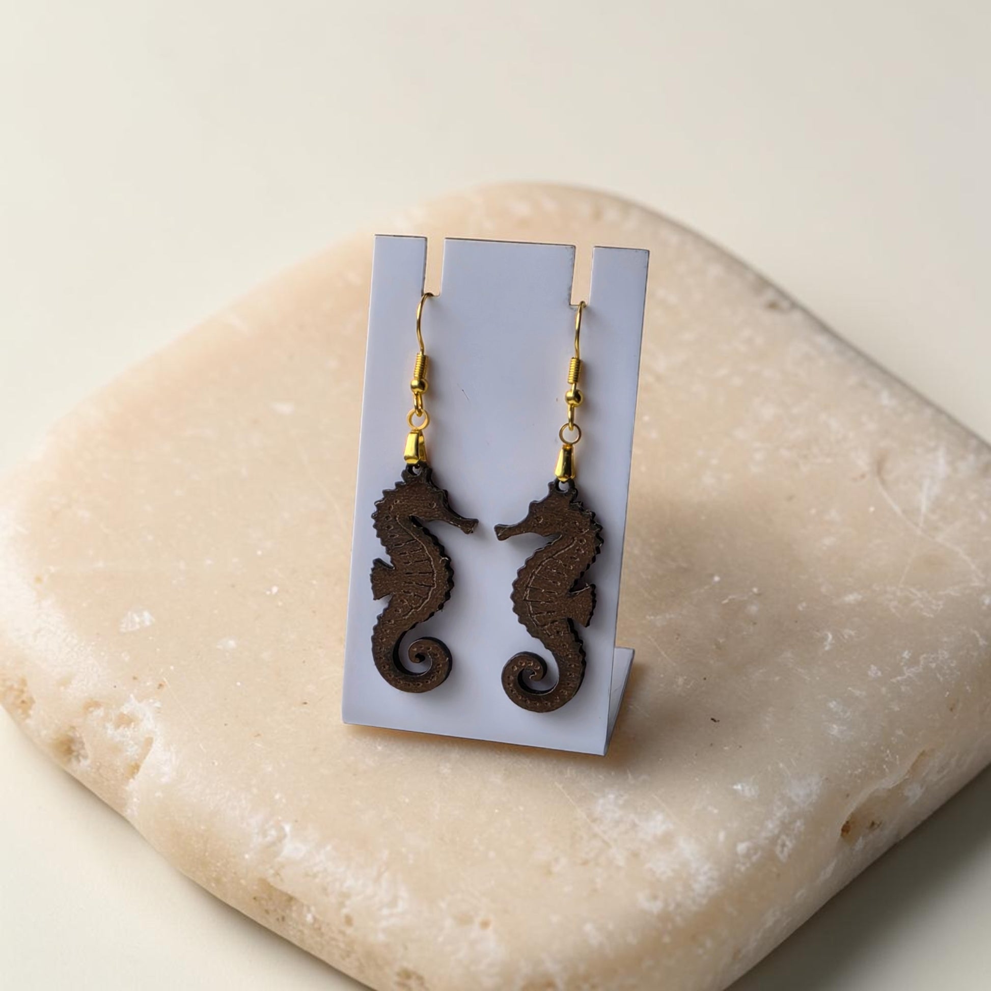 Seahorse Earrings | Lasercut Walnut Wood by Mirabilia Boutique—unique ocean jewelry with gold hooks, shown on a white card atop a smooth beige stone.