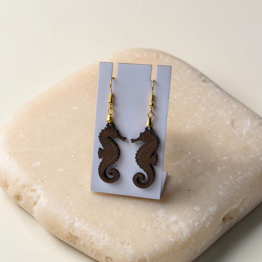 Seahorse Earrings | Lasercut Walnut Wood by Mirabilia Boutique—unique ocean jewelry with gold hooks, shown on a white card atop a smooth beige stone.