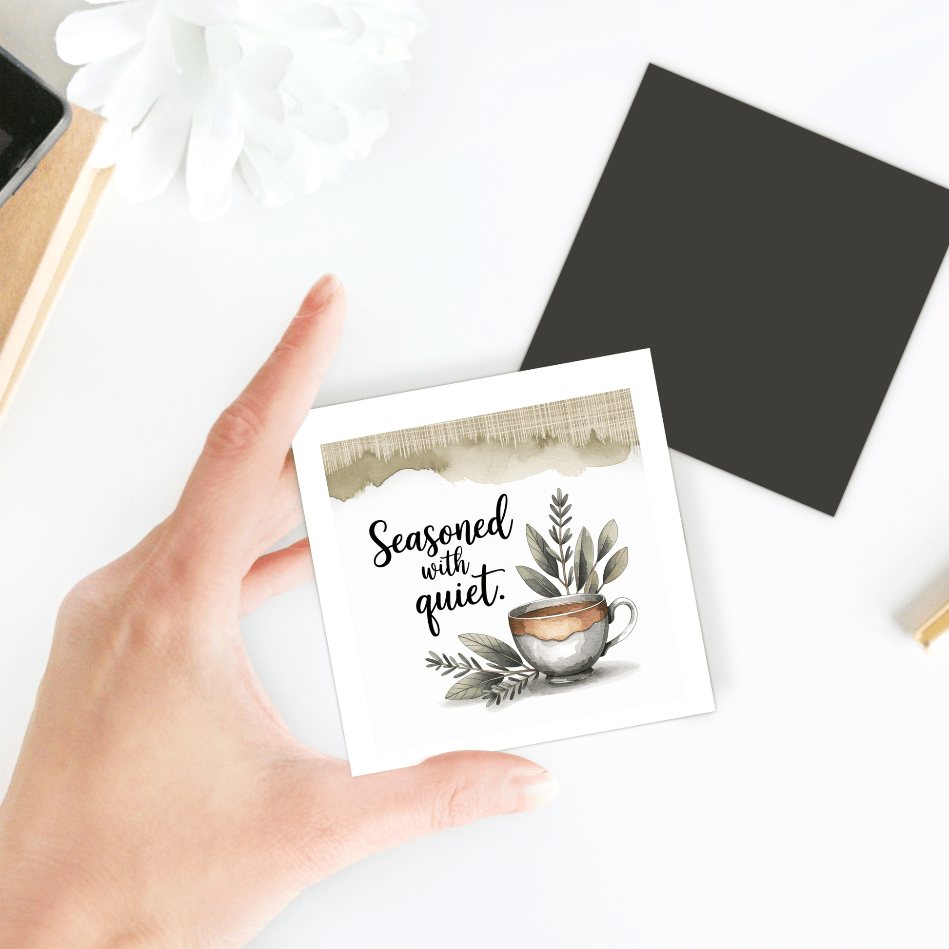 A hand holds the Mirabilia Boutique "Seasoned with Quiet Magnet | Calm Kitchen Decor Gift," featuring a coffee cup and green leaves—perfect for mindful spaces. A black card and white flower in the background create a serene vibe.