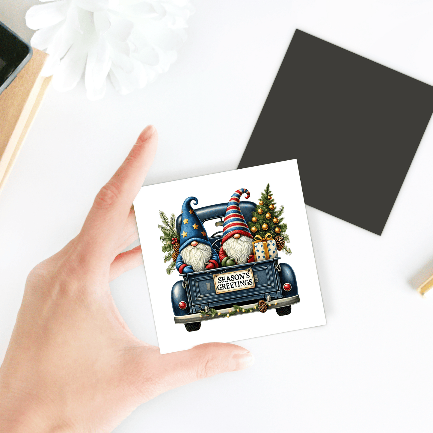 A hand holds the Season’s Greetings Gnome Magnet by Mirabilia Boutique, featuring two gnomes in a blue truck with gifts and a Christmas tree. A perfect holiday decor piece, shown on a white surface with a white flower and black square in the background.