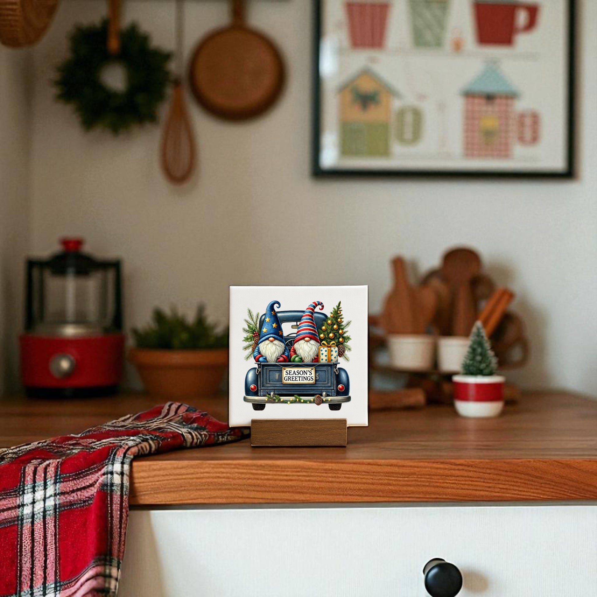 The Season’s Greetings Gnome Mini Sign by Mirabilia Boutique, a 4x4 holiday canvas, sits on a wooden kitchen counter with festive decor and a red plaid cloth, creating a charming Christmas tabletop display.