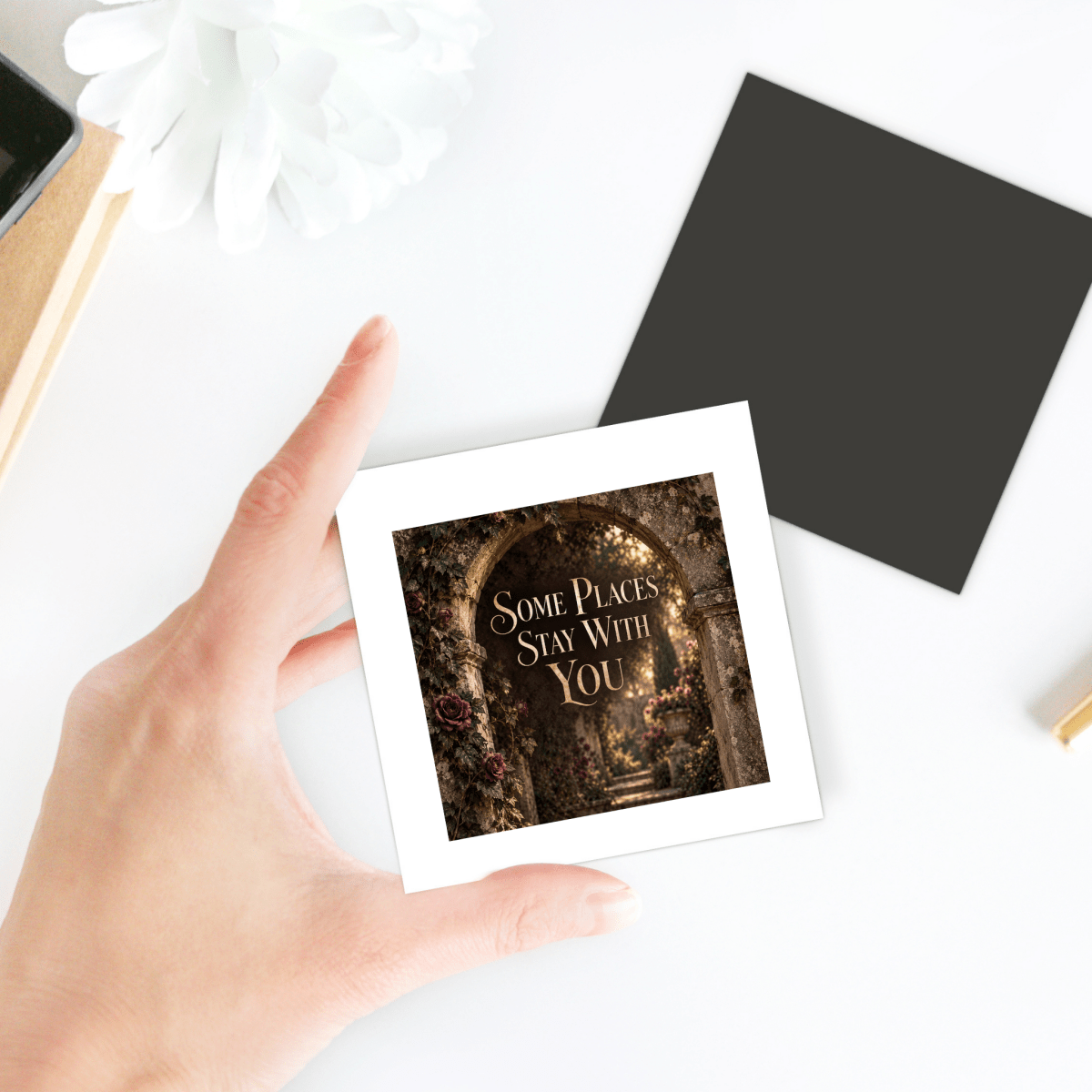 A hand holds the Some Places Stay With You Magnet from Mirabilia Boutique, featuring an image of an archway and flowers with the words “Some Places Stay With You.” Perfect as a thoughtful gift, this Velvet Ruins decorative magnet is shown alongside a blank black square and a white flower on the white tabletop.