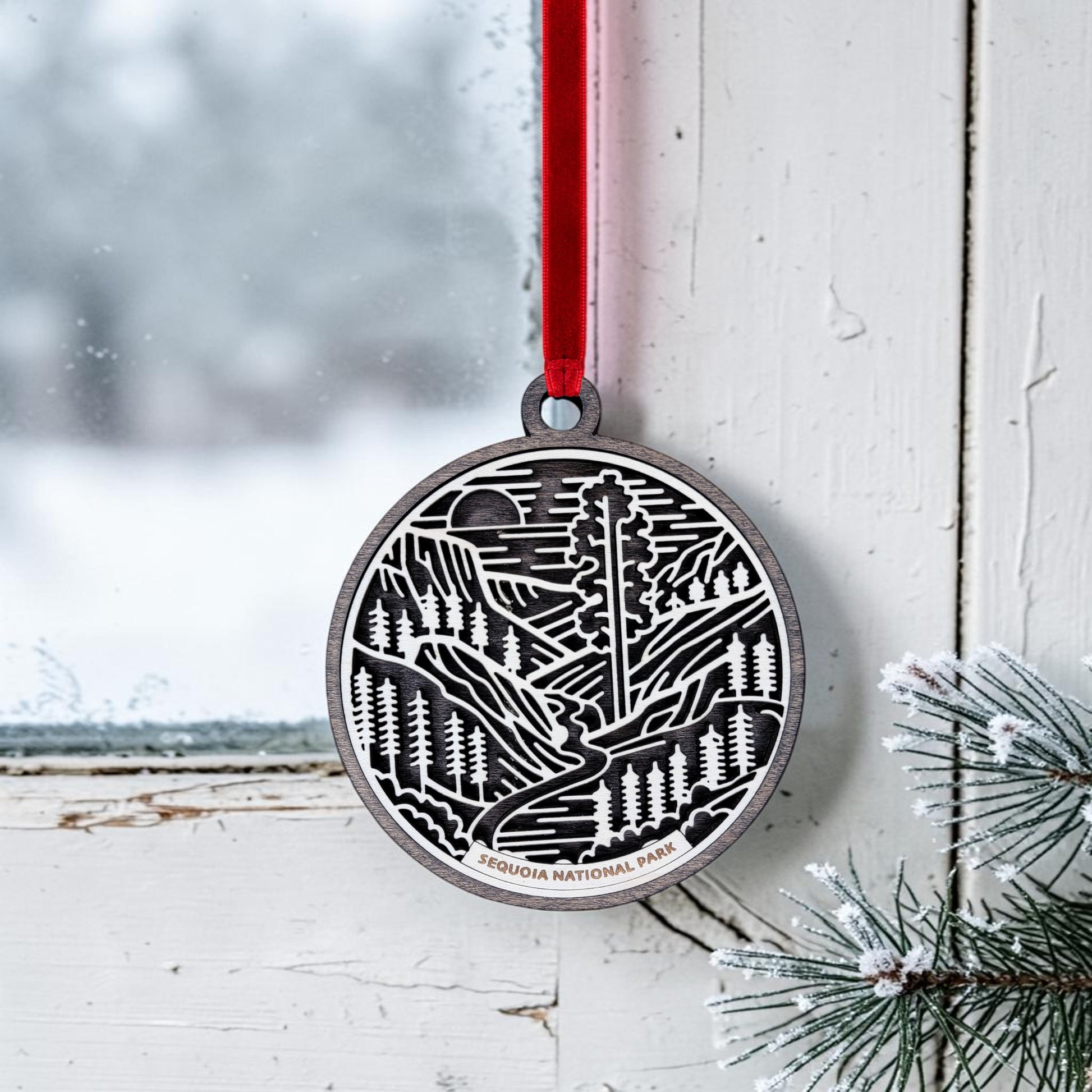 A round metal Sequoia National Park Line Art Christmas Ornament by Mirabilia Boutique, featuring an engraved forest and mountain scene, hangs by a red ribbon on a snowy window. Snow-dusted pine branches appear in the corner. The ornament’s text reads "Sequoia National Park," making this giant sequoia keepsake a perfect tribute to nature’s beauty.