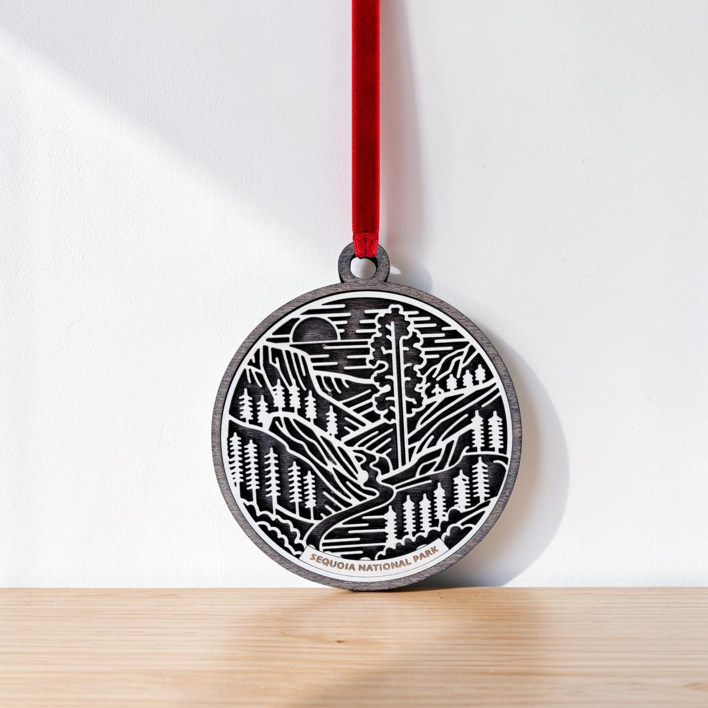 A round metal Sequoia National Park Line Art Christmas Ornament by Mirabilia Boutique, featuring a forest and mountain design and labeled "Sequoia National Park," hangs by a red ribbon against a white wall and rests on a light wooden surface—an ideal choice for anyone who loves giant sequoia art.