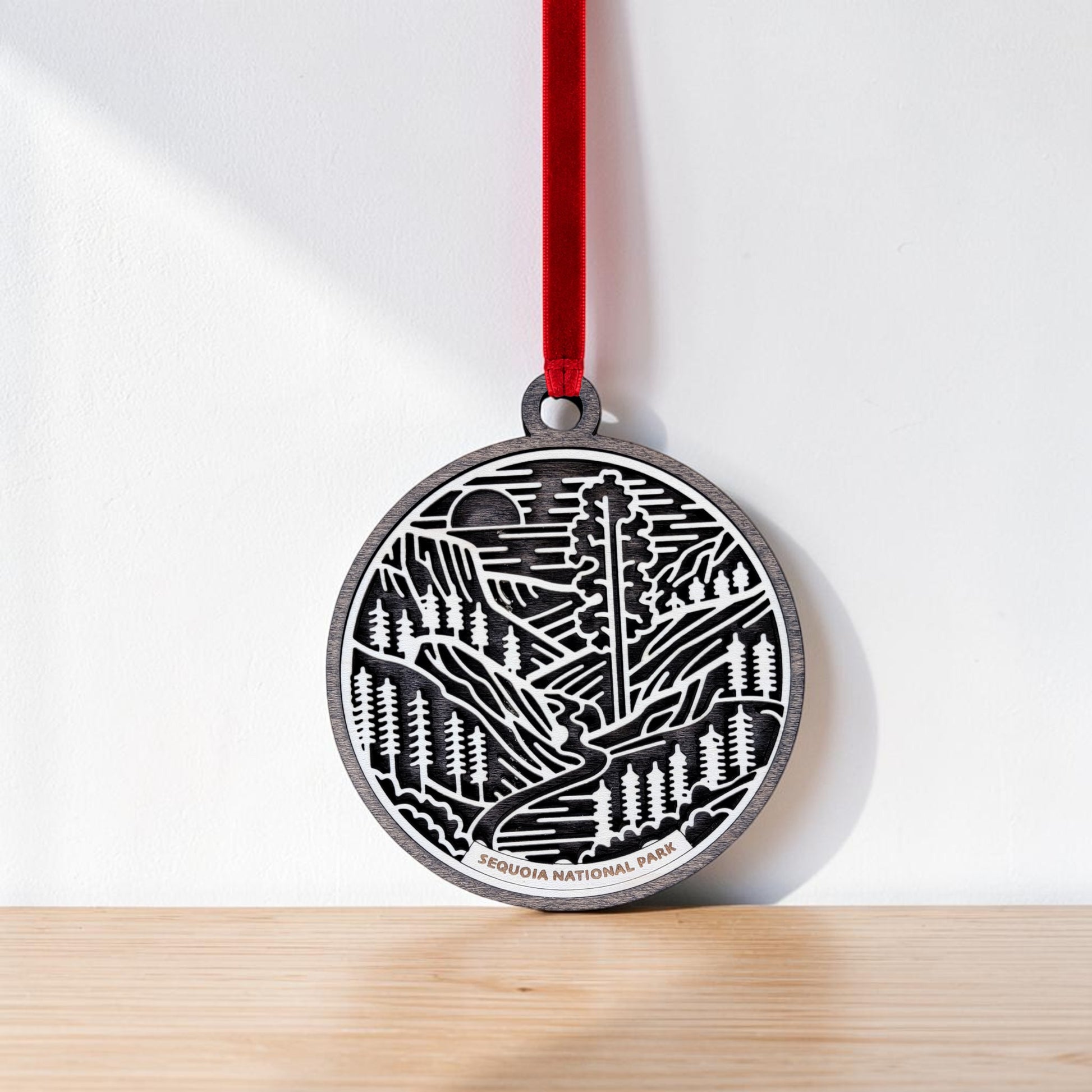 A round metal Sequoia National Park Line Art Christmas Ornament by Mirabilia Boutique, featuring a forest and mountain design and labeled "Sequoia National Park," hangs by a red ribbon against a white wall and rests on a light wooden surface—an ideal choice for anyone who loves giant sequoia art.