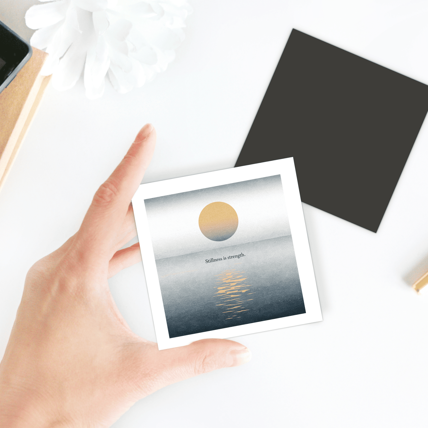 A hand holds the Mirabilia Boutique “Stillness Is Strength Magnet – Calm Minimalist Decor,” with golden sun art over water and inspiring text. Ideal for mindful gifting; background shows a black square, white flower, and desk items.