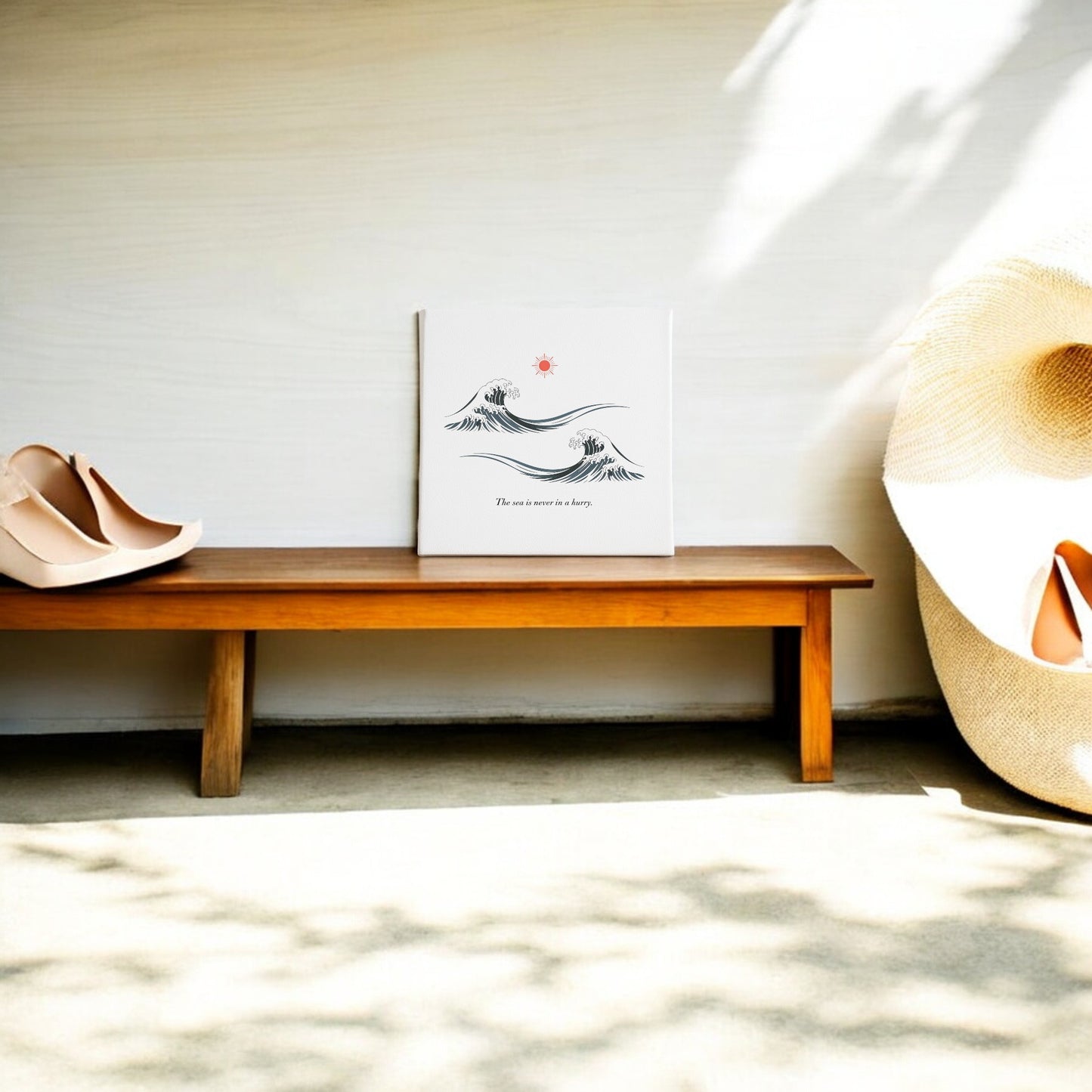 A wooden bench displays Mirabilia Boutique's "The Sea Is Never in a Hurry Canvas – Serene Ocean Calm," with a pair of nude shoes and a large woven hat. Sunlight streams in, casting tranquil patterns on the floor and wall.