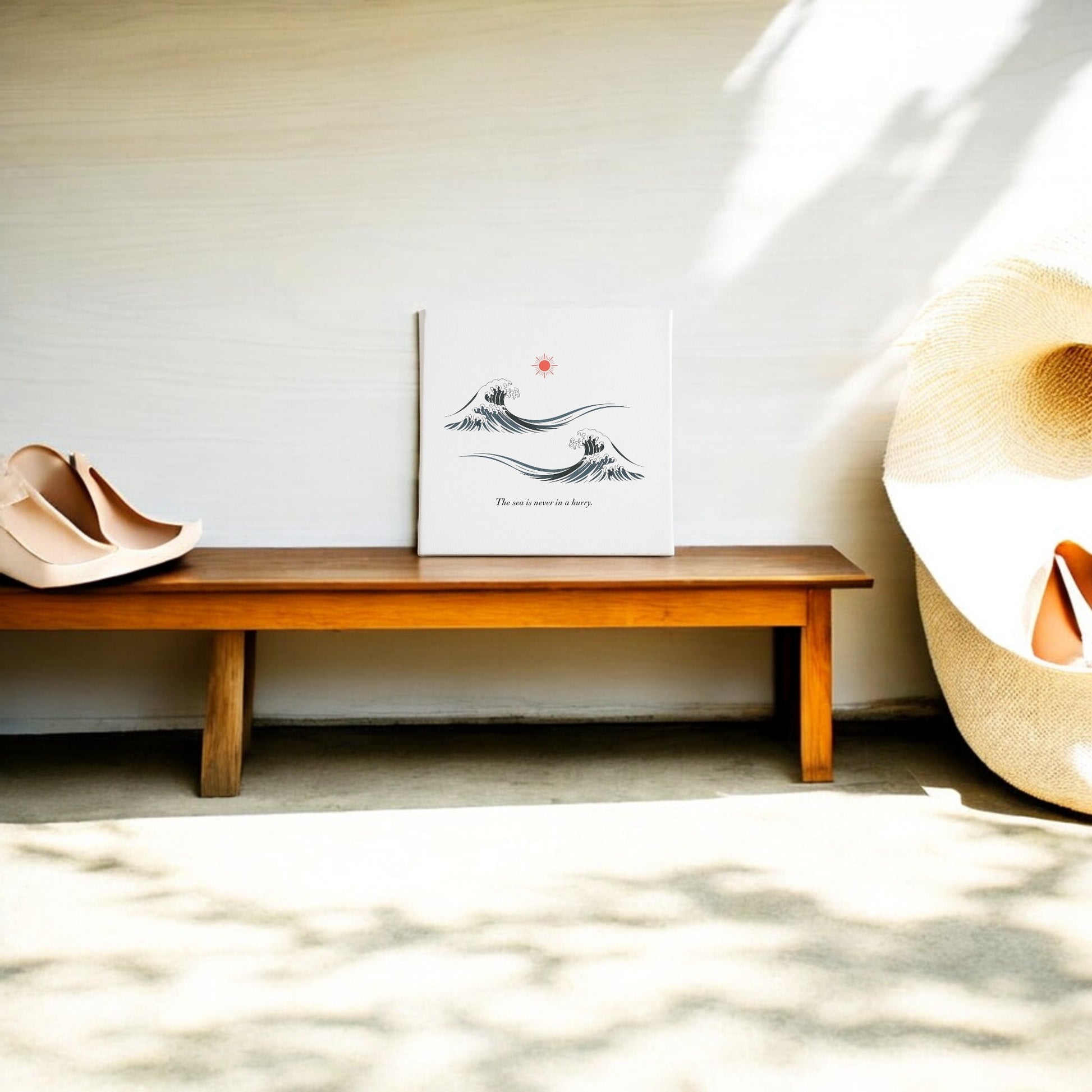 A wooden bench displays Mirabilia Boutique's "The Sea Is Never in a Hurry Canvas – Serene Ocean Calm," with a pair of nude shoes and a large woven hat. Sunlight streams in, casting tranquil patterns on the floor and wall.