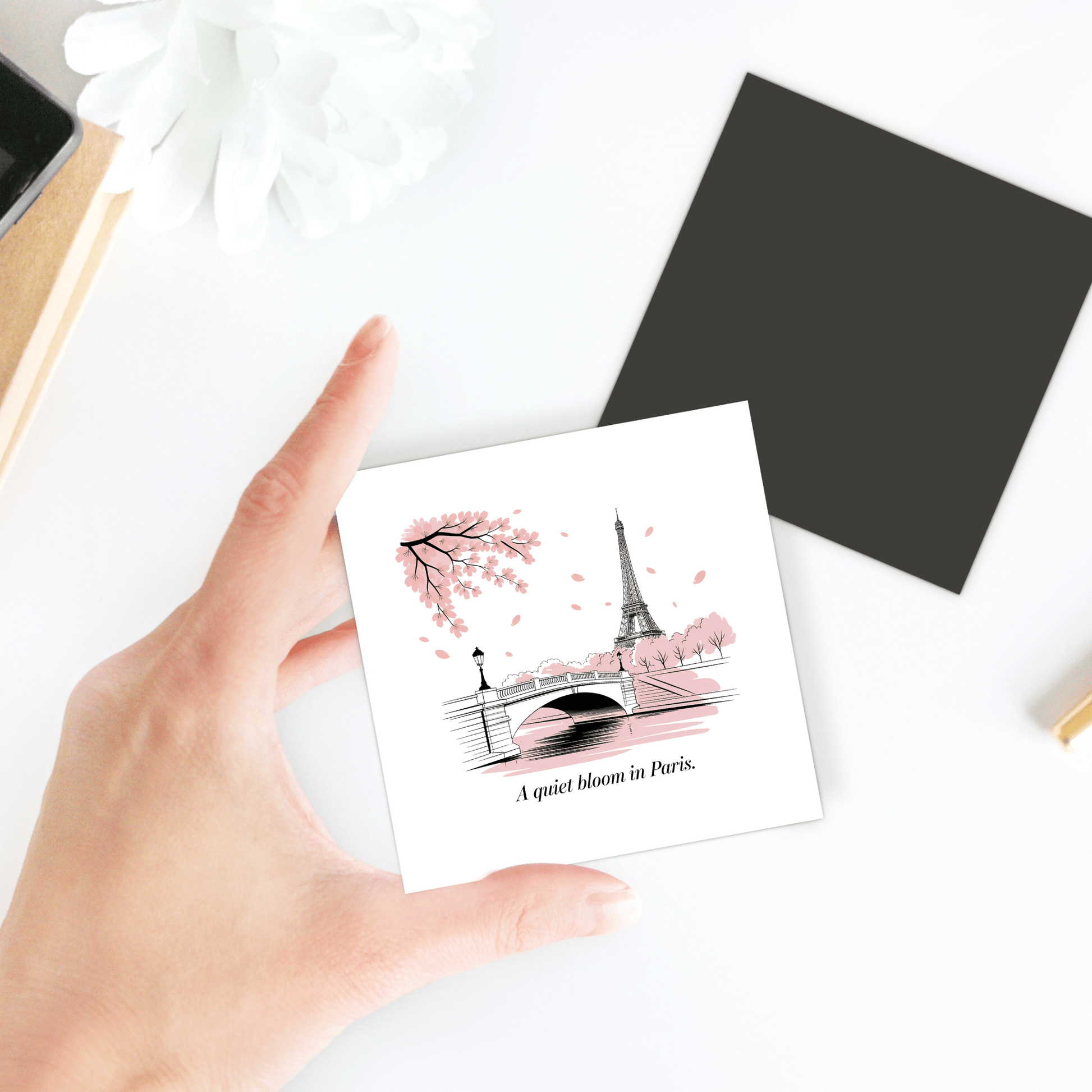A hand holds Mirabilia Boutique's "A Quiet Bloom in Paris – Cherry Blossom Magnet," featuring the Eiffel Tower, a bridge, and pink blooms. The product rests on a white table with a flower and black card.