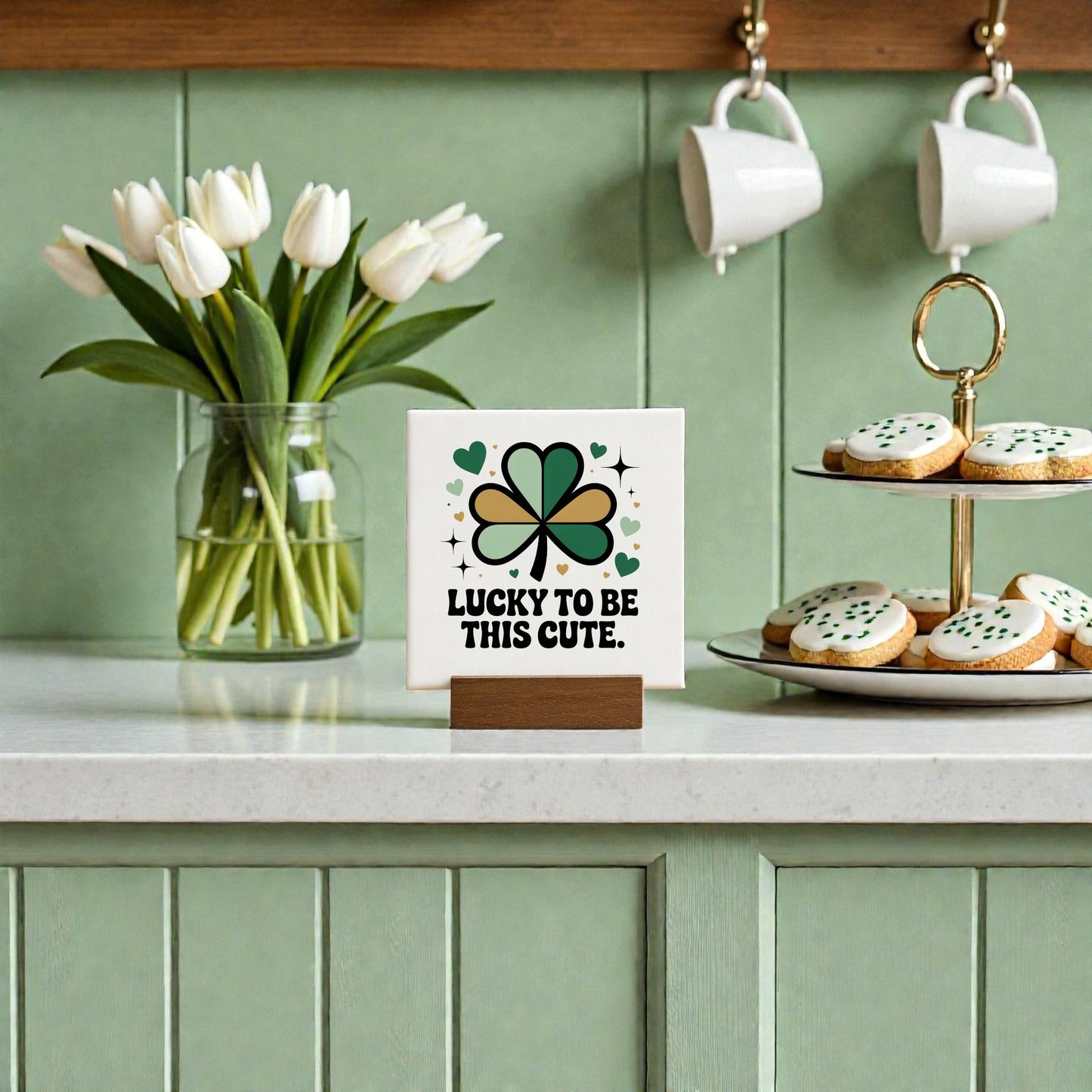 A kitchen scene with a Mirabilia Boutique "Lucky to Be This Cute" 4x4 canvas sign, a vase of white tulips, clover accents, and green and white iced cookies on a light countertop—perfect for St. Patrick’s Day gifting.