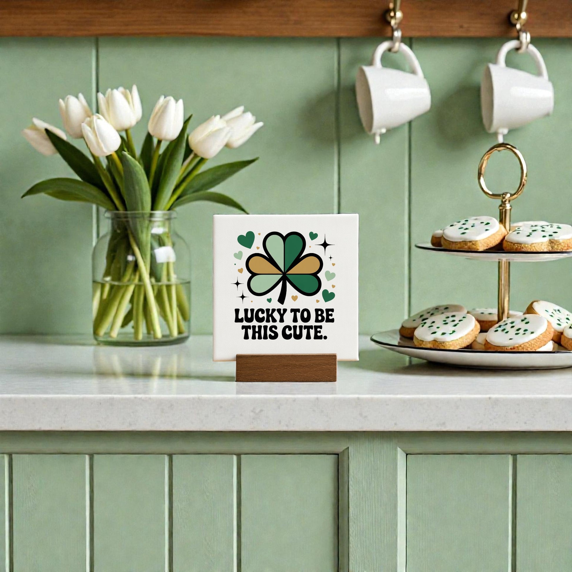 A kitchen scene with a Mirabilia Boutique "Lucky to Be This Cute" 4x4 canvas sign, a vase of white tulips, clover accents, and green and white iced cookies on a light countertop—perfect for St. Patrick’s Day gifting.