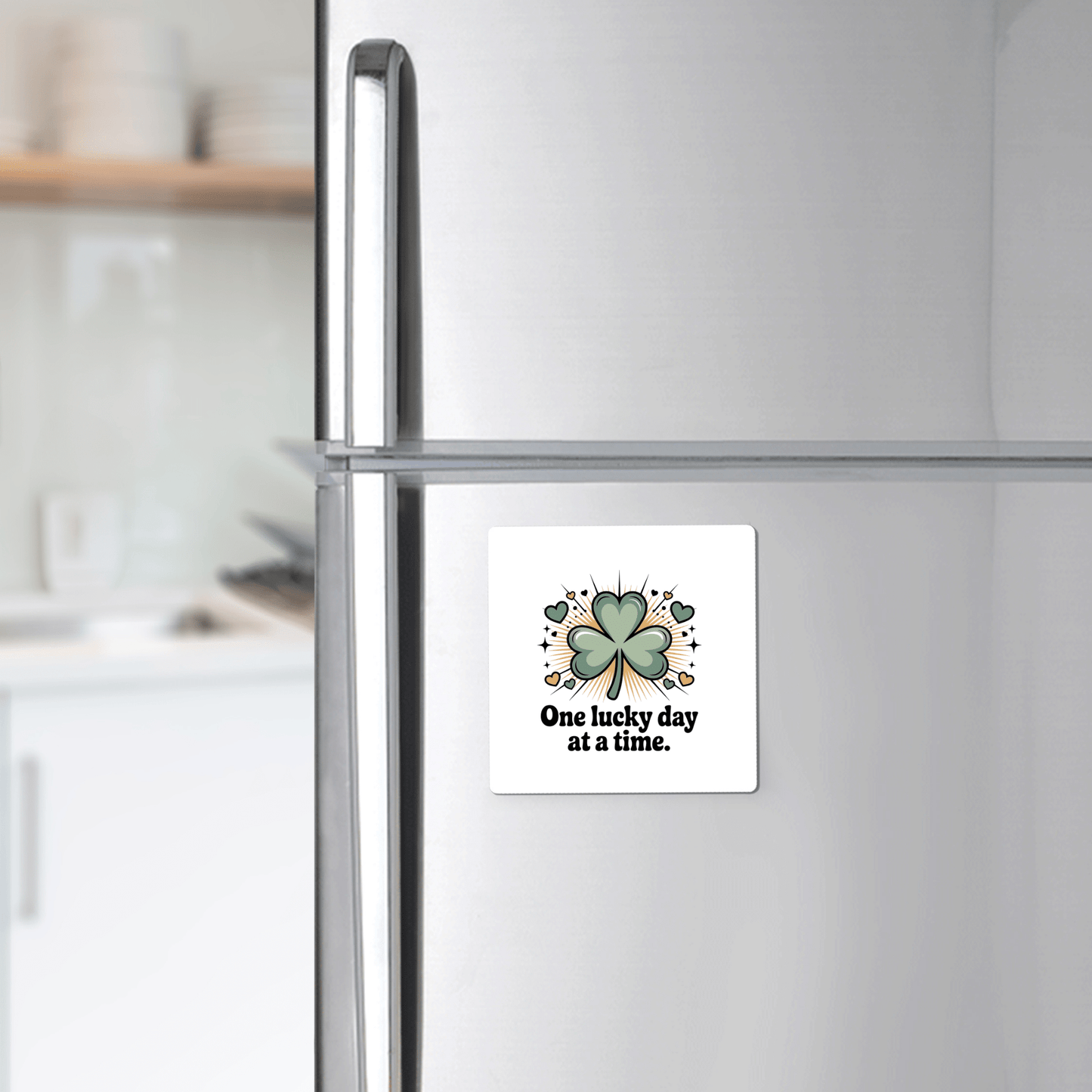 A Mirabilia Boutique "One Lucky Day at a Time Magnet" with a four-leaf clover decorates a refrigerator; kitchen counter and shelves with dishes appear blurred in the background.