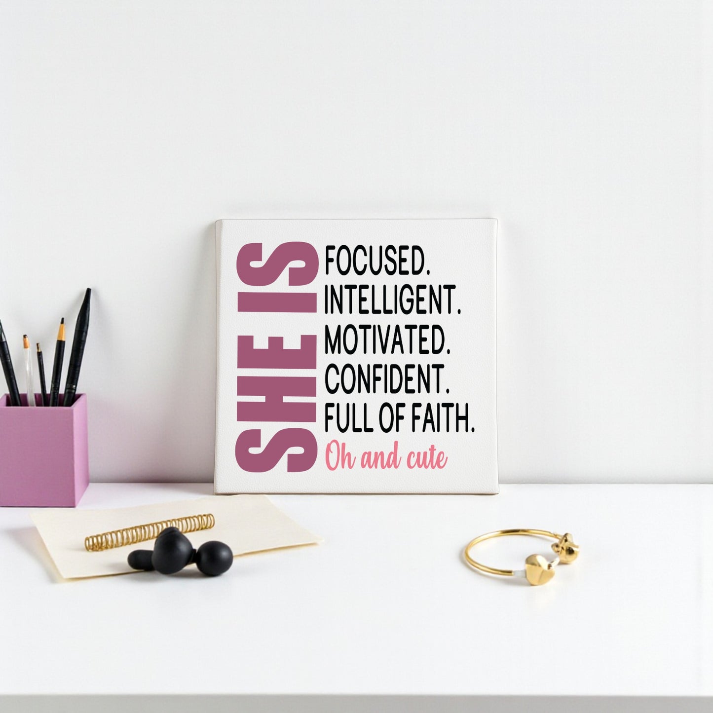 A white desk features the Mirabilia Boutique "She Is Focused Motivated Confident" 8x8 canvas wall art with affirmations, a pink pen holder, notebook, black beads, and a gold bangle arranged neatly on the surface.