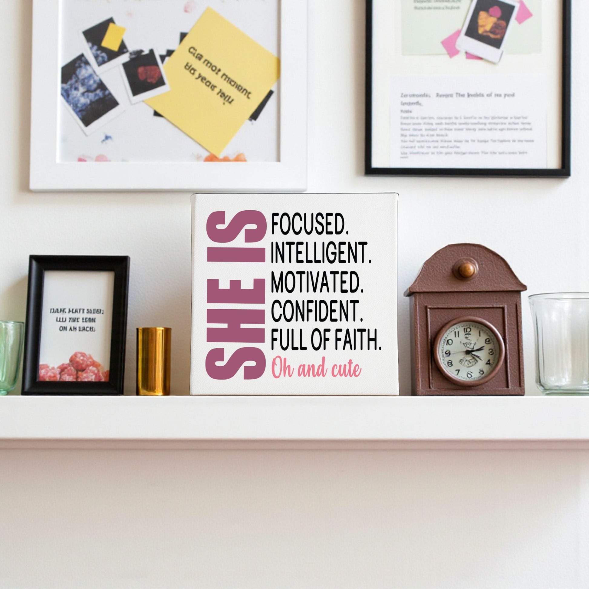 A white shelf displays framed photos, a small clock, and the Mirabilia Boutique "She Is Focused Motivated Confident" 8x8 canvas wall art, with empowering affirmations framed on the white wall in the background.