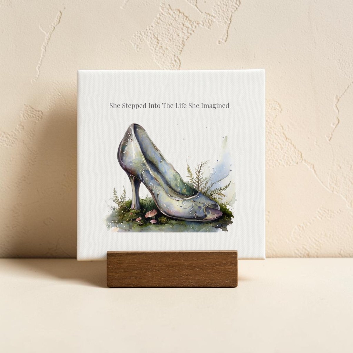 The She Stepped Into the Life She Imagined Mini Sign by Mirabilia Boutique features a whimsical watercolor high heel shoe, delicate greenery accents, and the inspiring phrase "She Stepped Into The Life She Imagined," beautifully displayed on a wooden stand against a light textured wall.