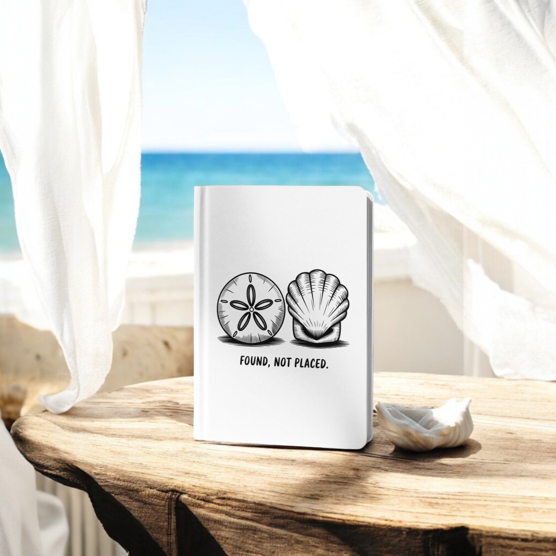 The Found, Not Placed Journal – Coastal Quote Notebook by Mirabilia Boutique, featuring seashell and sand dollar illustrations, sits on a rustic wood table by a window overlooking the ocean and blue sky.