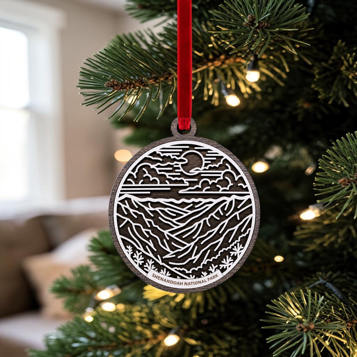 The Mirabilia Boutique Shenandoah National Park Line Art Christmas Ornament, handcrafted from wood and elegantly oval-shaped, hangs from a Christmas tree branch adorned with lights. Featuring detailed Blue Ridge Mountains artwork in a mountain and sky motif and accented by a vibrant red ribbon, this keepsake is the perfect tribute to your love of nature and adventure.