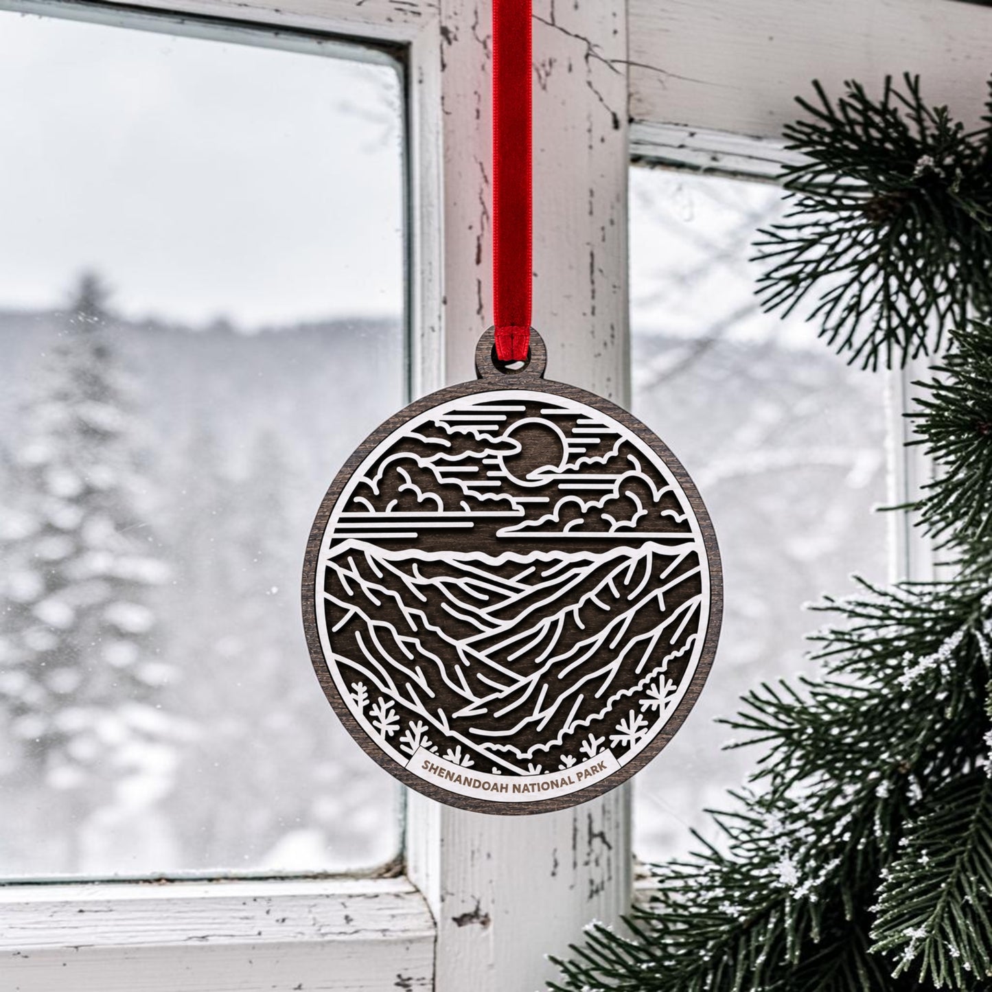 A handcrafted wooden Shenandoah National Park Line Art Christmas Ornament by Mirabilia Boutique, featuring a mountain and sun design and labeled "Shenandoah National Park," hangs by a red ribbon in front of a snowy window beside evergreen branches. This decorative piece beautifully captures the essence of the Blue Ridge Mountains, making it a perfect addition to your holiday décor.