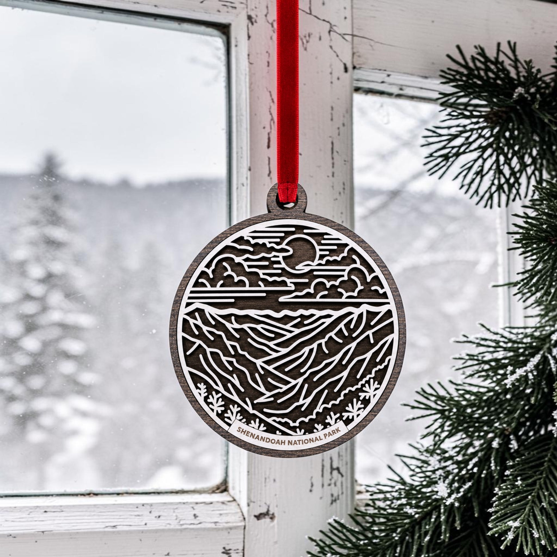 A handcrafted wooden Shenandoah National Park Line Art Christmas Ornament by Mirabilia Boutique, featuring a mountain and sun design and labeled "Shenandoah National Park," hangs by a red ribbon in front of a snowy window beside evergreen branches. This decorative piece beautifully captures the essence of the Blue Ridge Mountains, making it a perfect addition to your holiday décor.