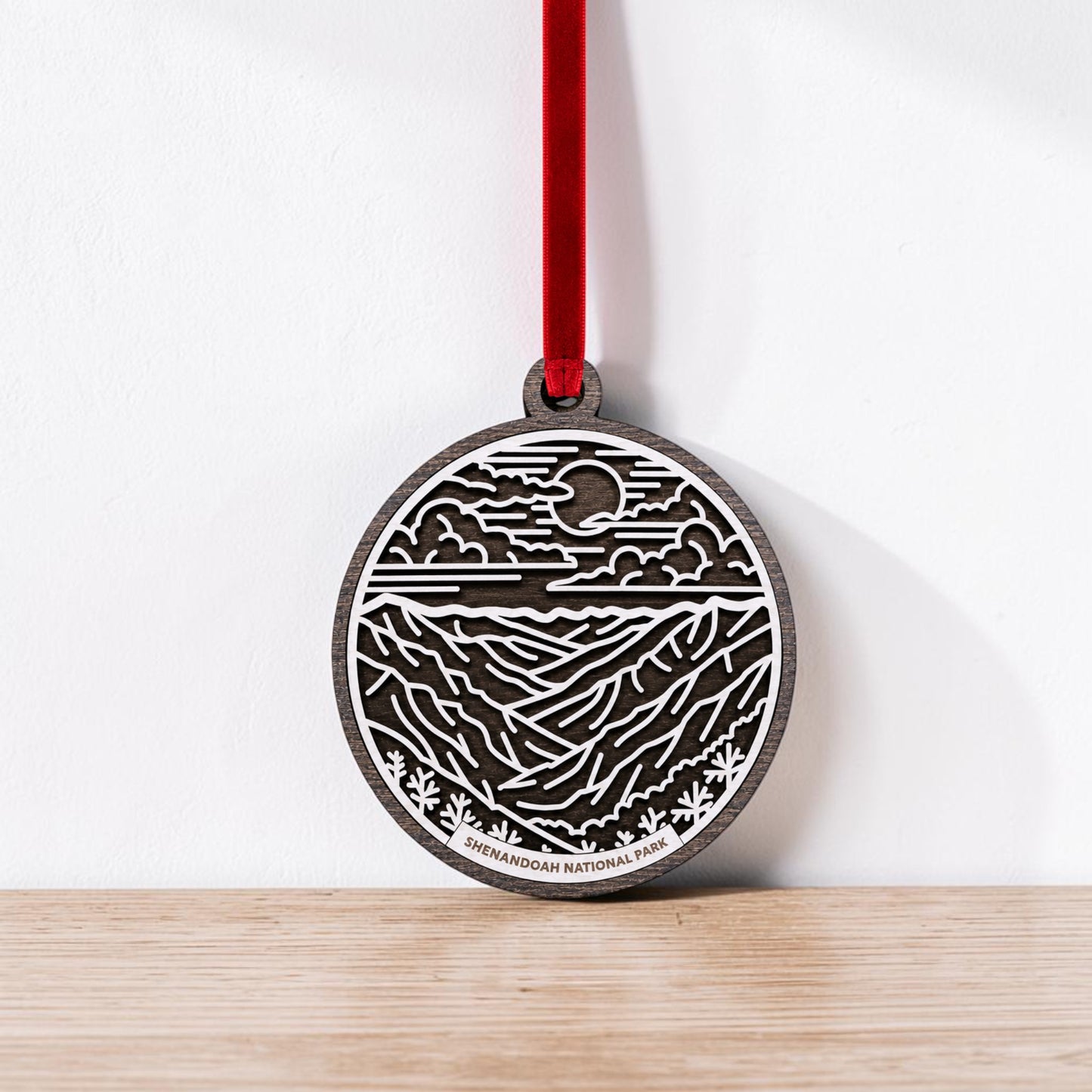The Shenandoah National Park Line Art Christmas Ornament by Mirabilia Boutique is a round, dark wood keepsake featuring a detailed mountain and sky scene in white. Suspended from a red ribbon, it displays "Shenandoah National Park" at the bottom and rests against a white wall.