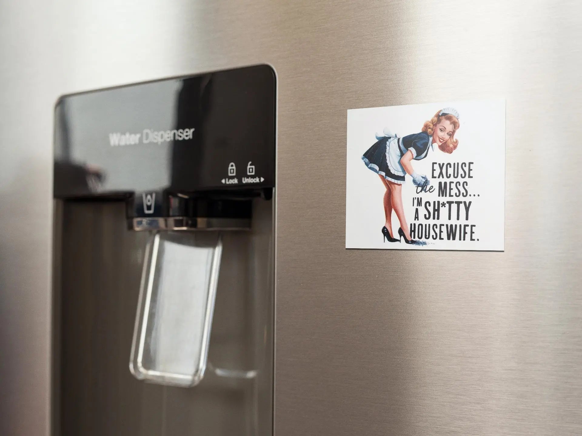 The stainless steel refrigerator with a built-in water dispenser is adorned with the Retro "Excuse the Mess" Shitty Housewife Magnet from Mirabilia Boutique, adding humorous fridge decor featuring a woman declaring, "Excuse the mess... I'm a shitty housewife." This quirky kitchen accessory brings charm and laughter to your culinary space.