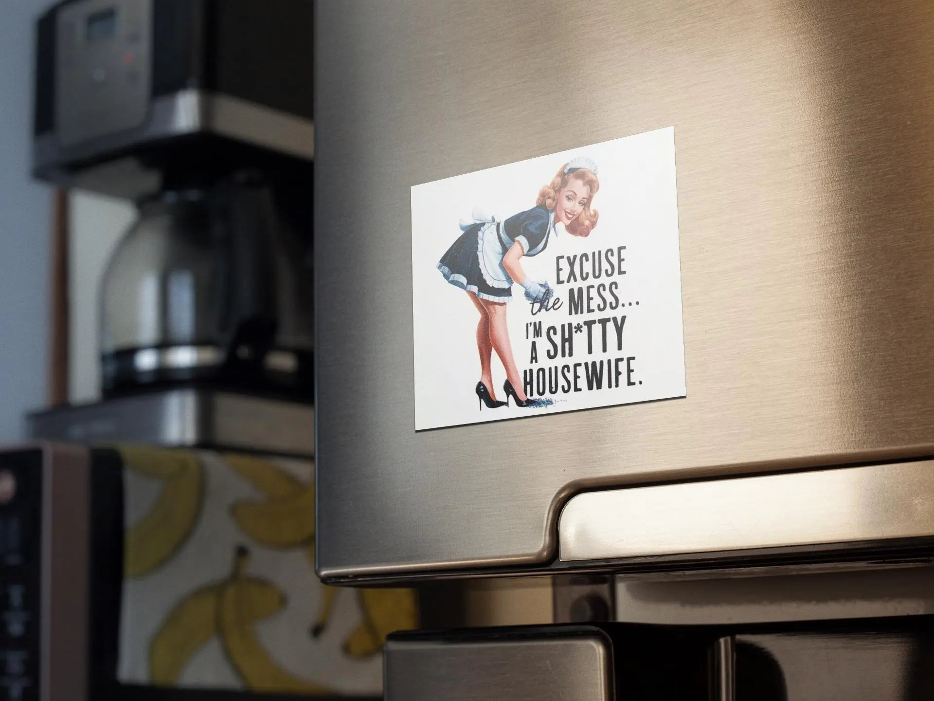 A refrigerator door is adorned with the Mirabilia Boutique's Retro "Excuse the Mess" Shitty Housewife Magnet, which humorously features a woman in a maid outfit bending over along with the text: "Excuse the mess... I'm a shitty housewife." This quirky kitchen accessory perfectly complements the coffee maker in the background.