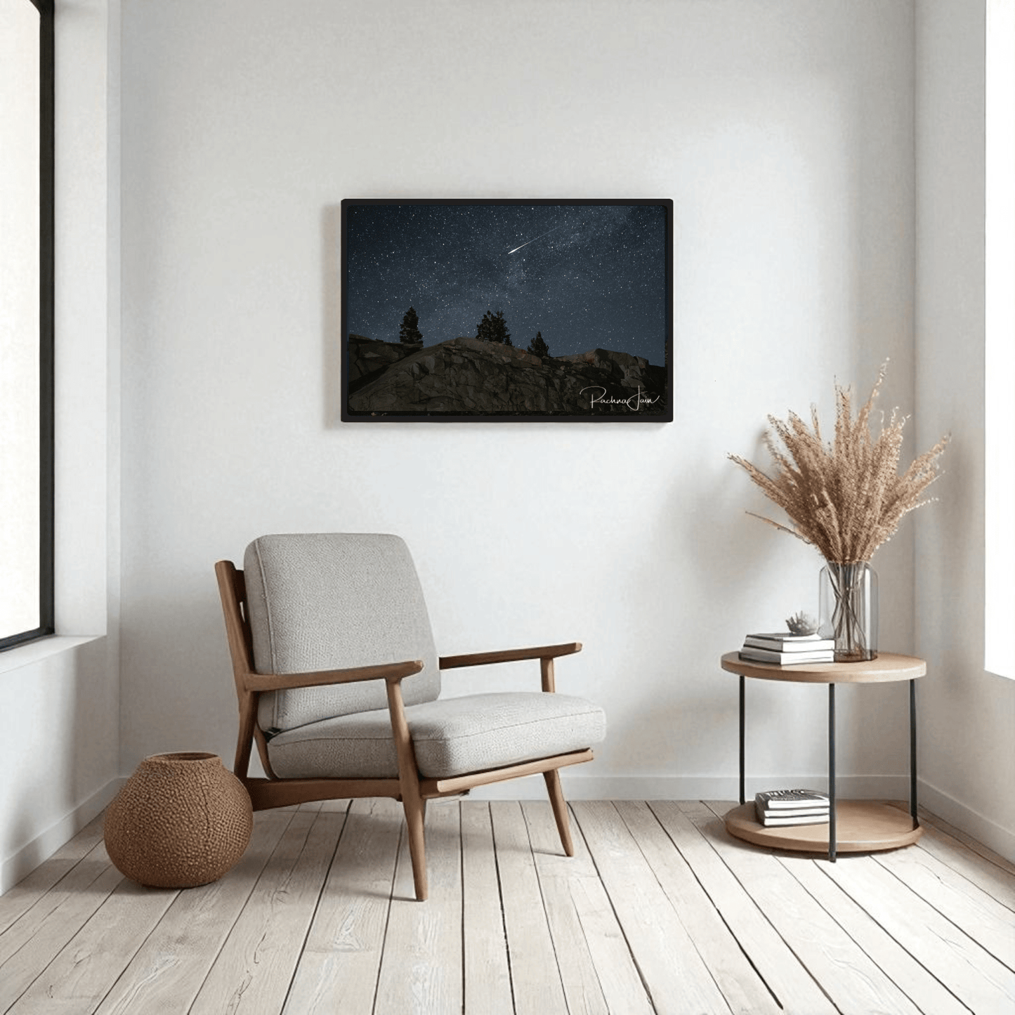 A cozy, modern living room with a light gray armchair, round wooden side table with a vase of dried grass, and Mirabilia Boutique’s "Shooting Star at Olmsted Point | Night Sky Framed Art Print" displayed above on the white wall.