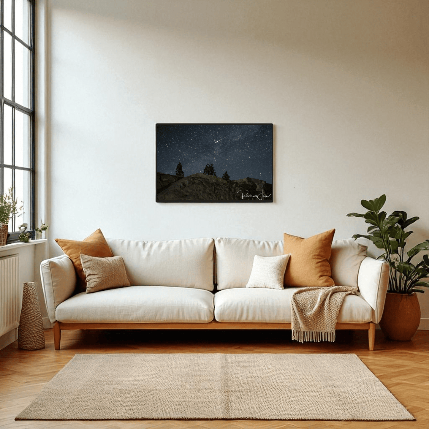 A cozy living room with a white sofa, tan cushions, potted plants, and a large window features Mirabilia Boutique’s "Shooting Star at Olmsted Point | Night Sky Framed Art Print" above the sofa, adding celestial charm to the inviting space.