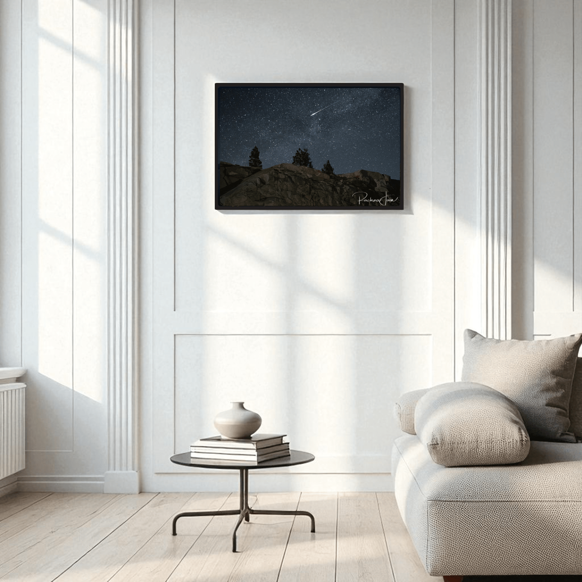 A modern living room with white walls, a light gray sofa, a round black coffee table with a vase, and "Shooting Star at Olmsted Point | Night Sky Framed Art Print" by Mirabilia Boutique above the sofa. Natural light streams through tall windows.