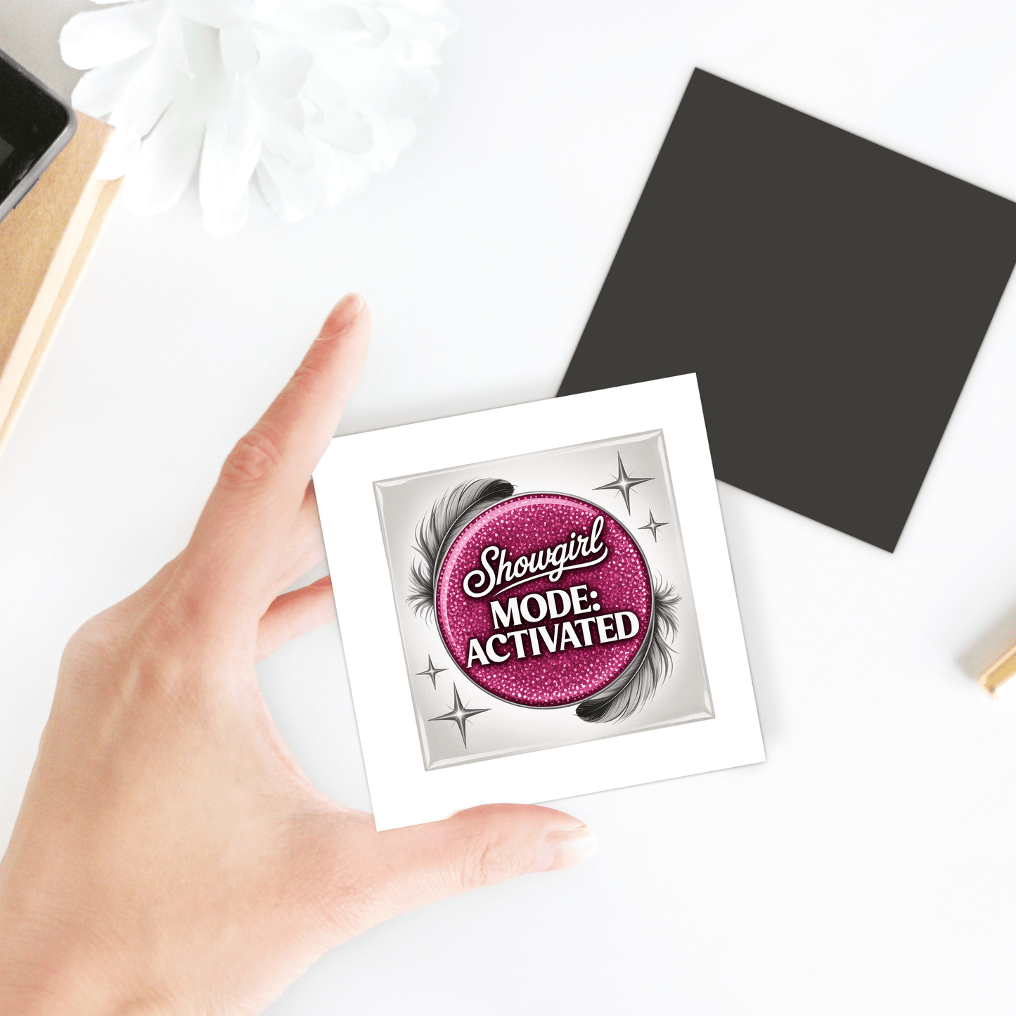 A hand holds the Mirabilia Boutique "Showgirl Mode Activated Magnet"—a glam pink glitter fridge decor with a sparkling design. Nearby are office items on a white surface. Perfect as a confidence gift.