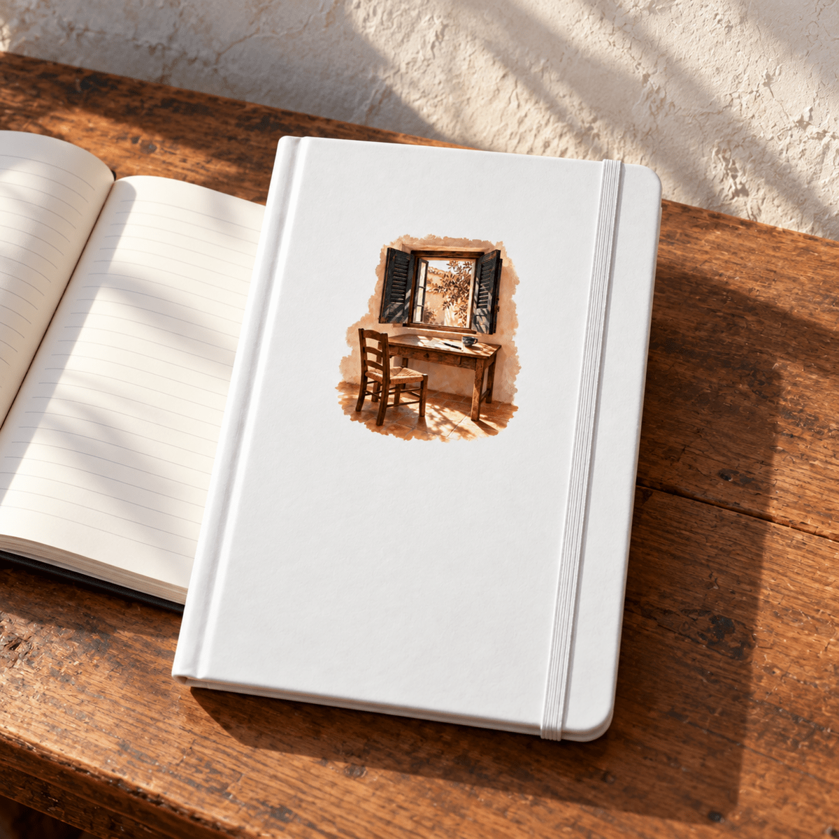 A white A5 The Shuttered Room Journal with an elastic closure sits on a wooden surface next to an open, lined notebook featuring a watercolor illustration of a desk by an open window.