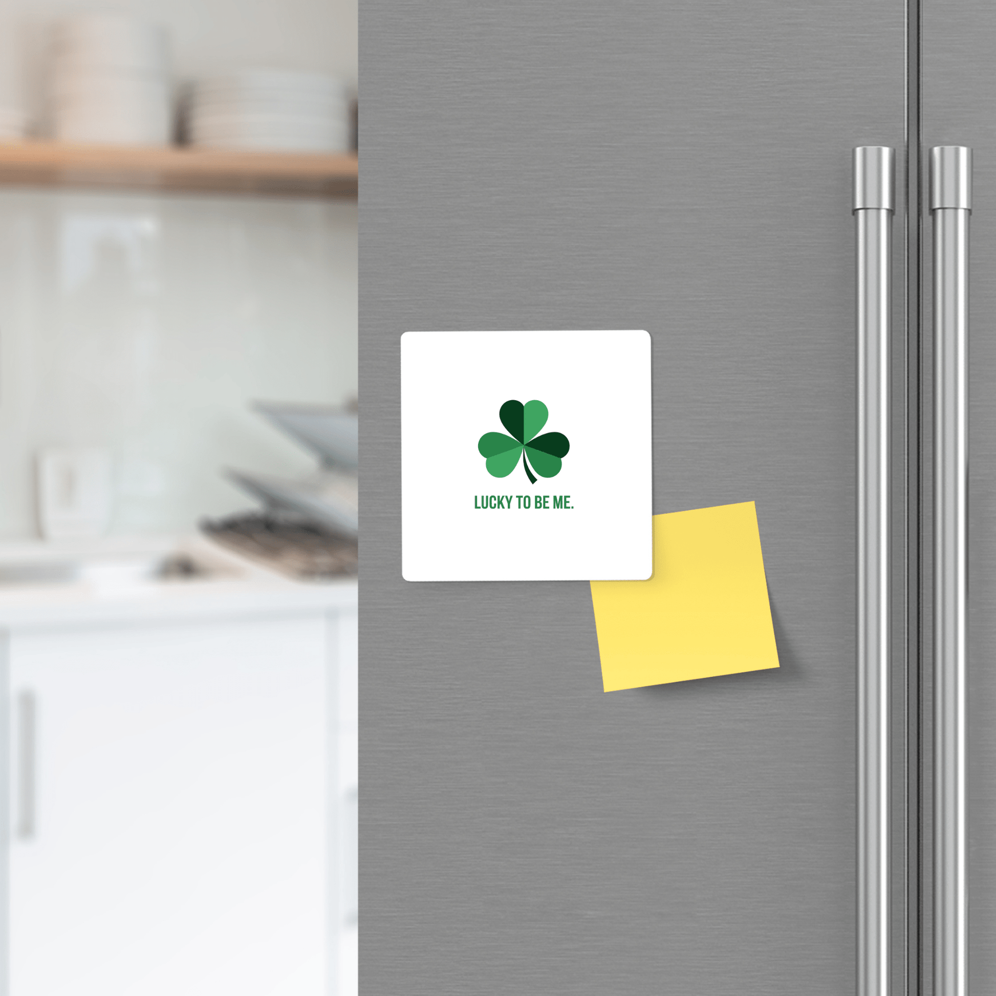 A modern kitchen features a refrigerator door adorned with Mirabilia Boutique's "Lucky to Be Me Shamrock Magnet" and a blank yellow sticky note beneath it.