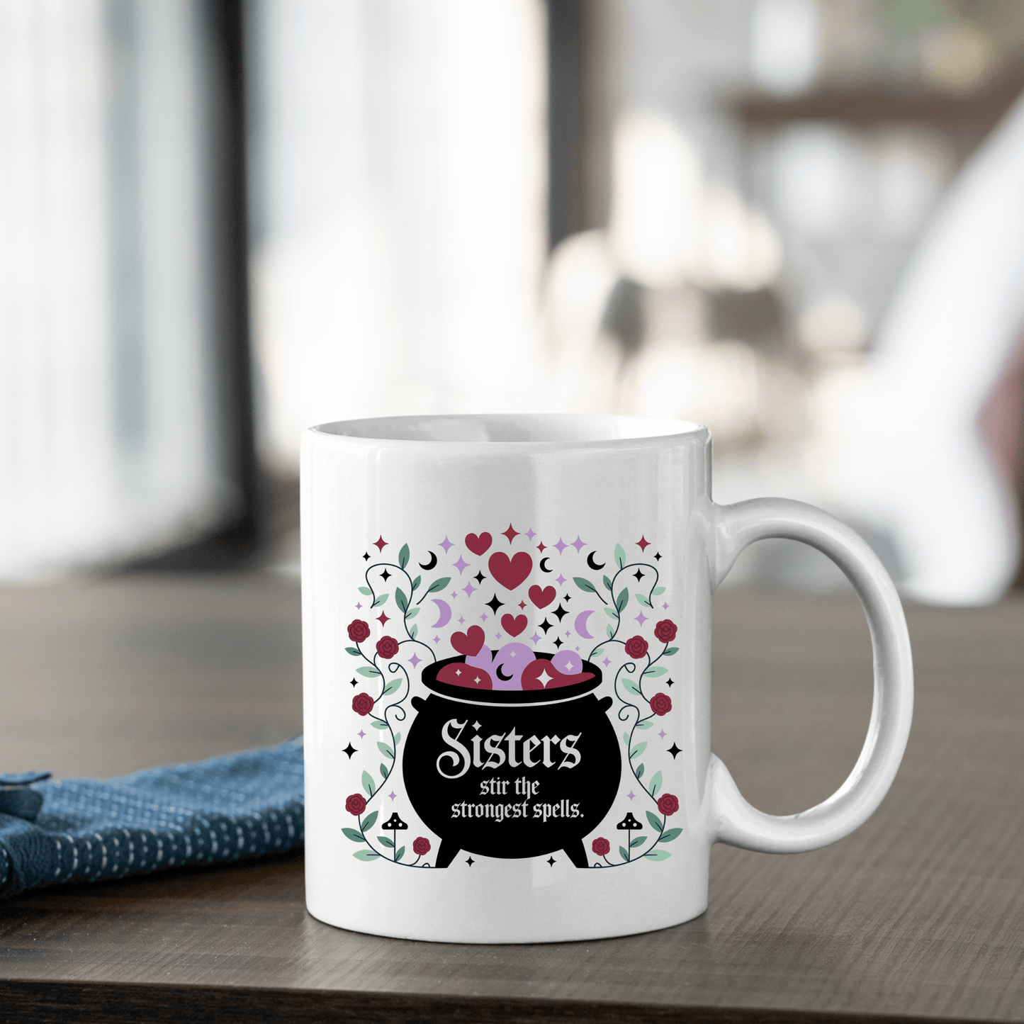 The Sisters Stir the Strongest Spells Mug by Mirabilia Boutique, featuring a black cauldron with pink hearts and flowers, is the perfect witchy sister gift—shown on a wooden table with a blue napkin.