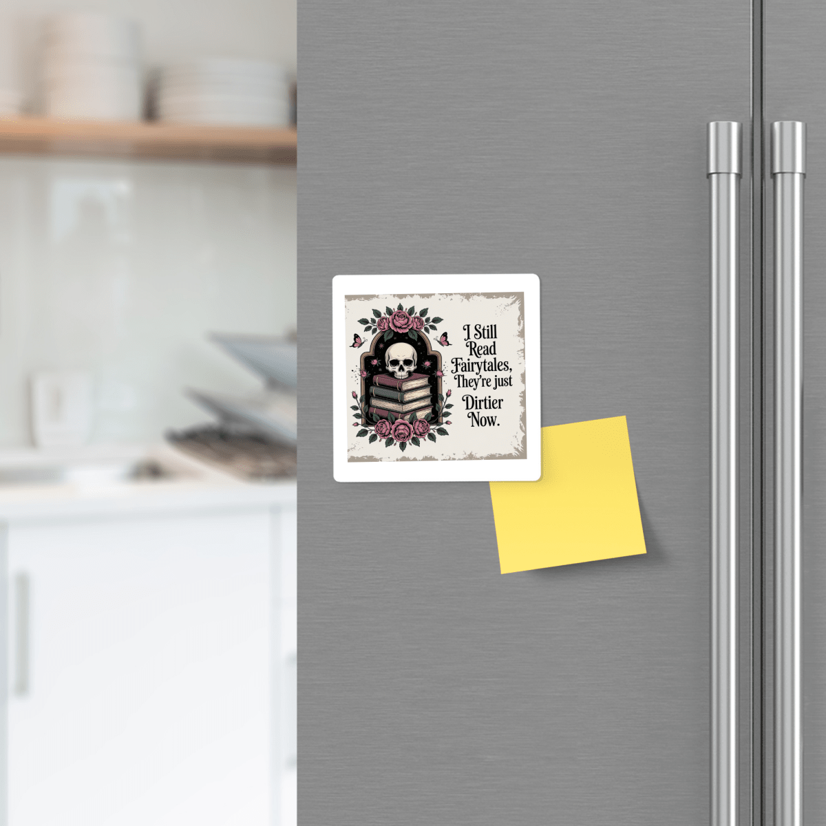 On a refrigerator door, the I Still Read Fairytales Gothic Book Magnet by Mirabilia Boutique—featuring a striking design of a skull, books, roses, and text—is displayed beside a yellow sticky note. Kitchen shelves with dishes appear in the background, highlighting this magnet as a perfect literary gift for fans of fairytale-inspired decor.