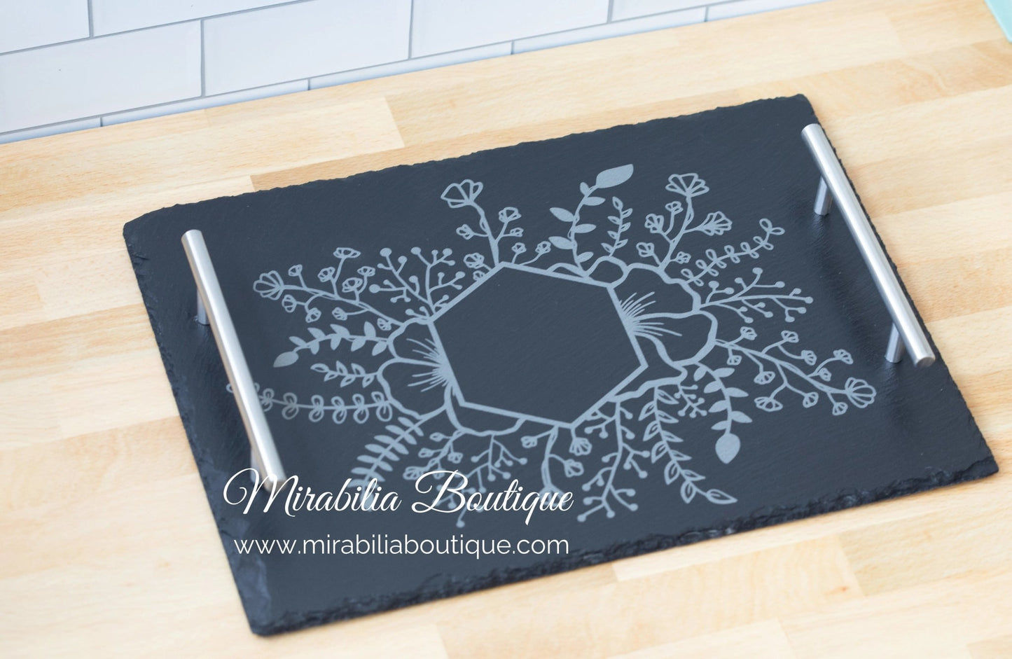The Personalized Engraved Slate Serving Tray with Handles by Mirabilia Boutique features a floral monogram design etched in the center, displayed on a light wooden countertop. "Mirabilia Boutique" and its website appear on the image.