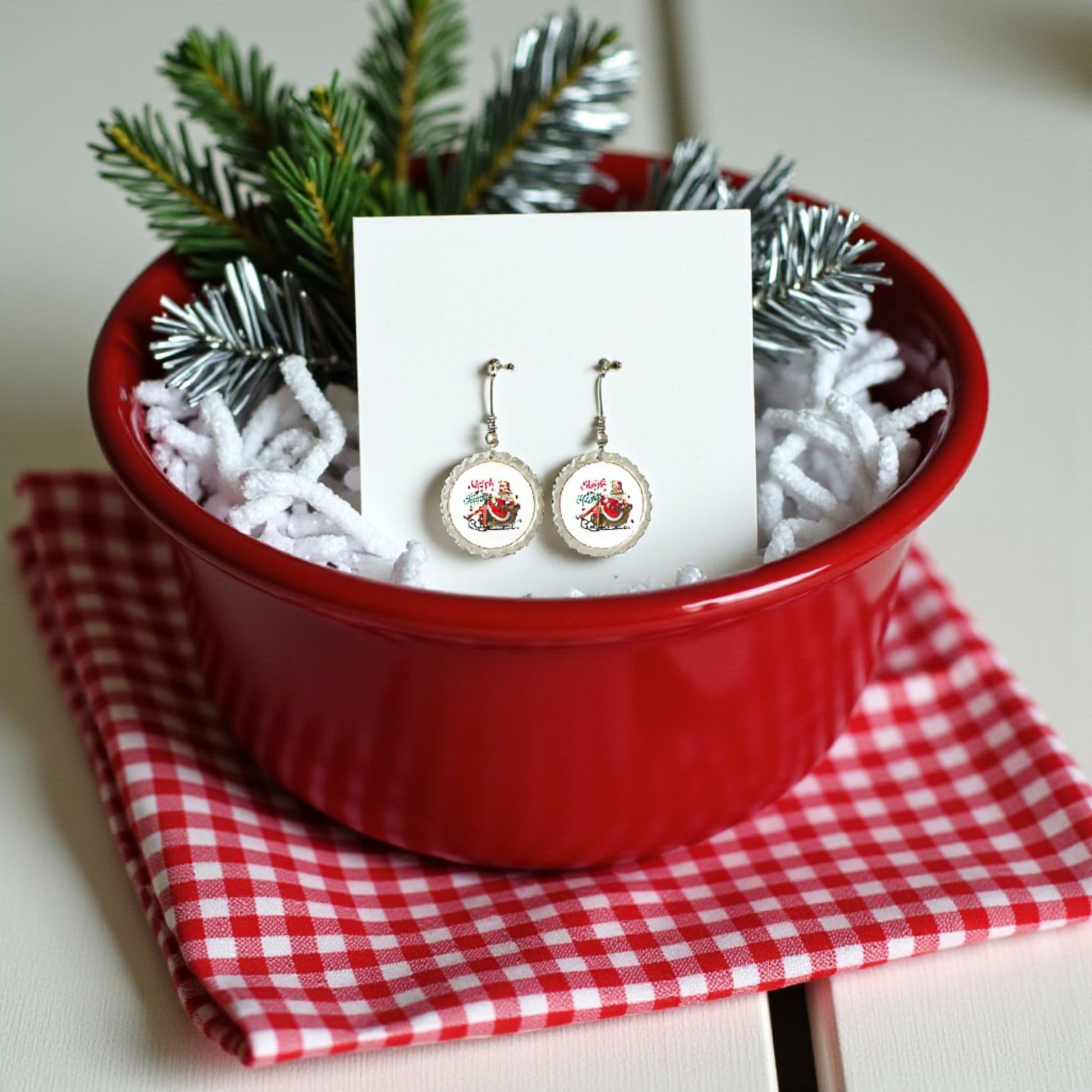 The Sleigh Girl Sleigh Earrings by Mirabilia Boutique feature festive red cardinals and are displayed on a card in a red bowl with white filler and pine branches atop a gingham cloth—your perfect retro holiday jewelry accent.