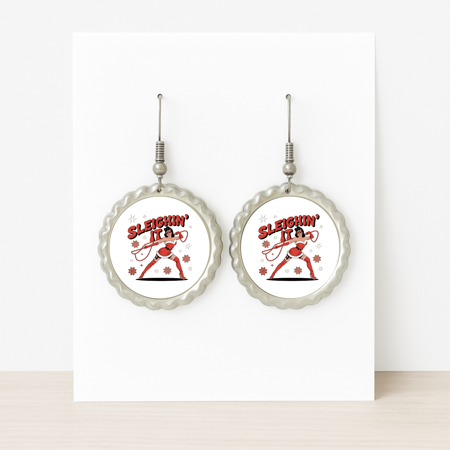 The Sleighin' It Earrings by Mirabilia Boutique are round, silver holiday earrings featuring a dancing Santa and "Sleighin' It" text in red—fun Christmas accessories for women, shown on a white background.