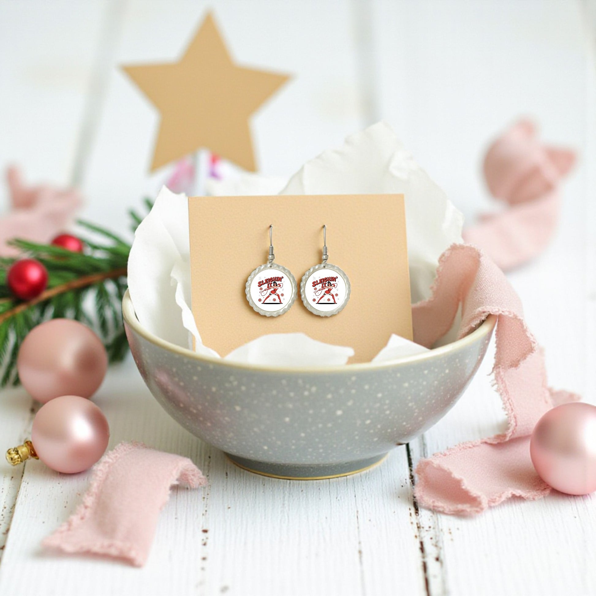 The Sleighin' It Earrings by Mirabilia Boutique are festive round holiday earrings displayed in a gray bowl with tissue paper, pink ornaments, ribbon, pine, and a gold star—perfect Christmas party accessories.