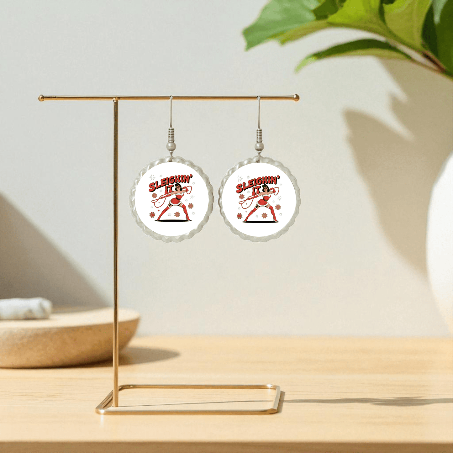 The Sleighin' It Earrings by Mirabilia Boutique feature white backgrounds, red "Sleighin' It" text, and a sleigh graphic. Displayed on a gold stand with a green plant behind, these fun holiday earrings are perfect Christmas party accessories for women.