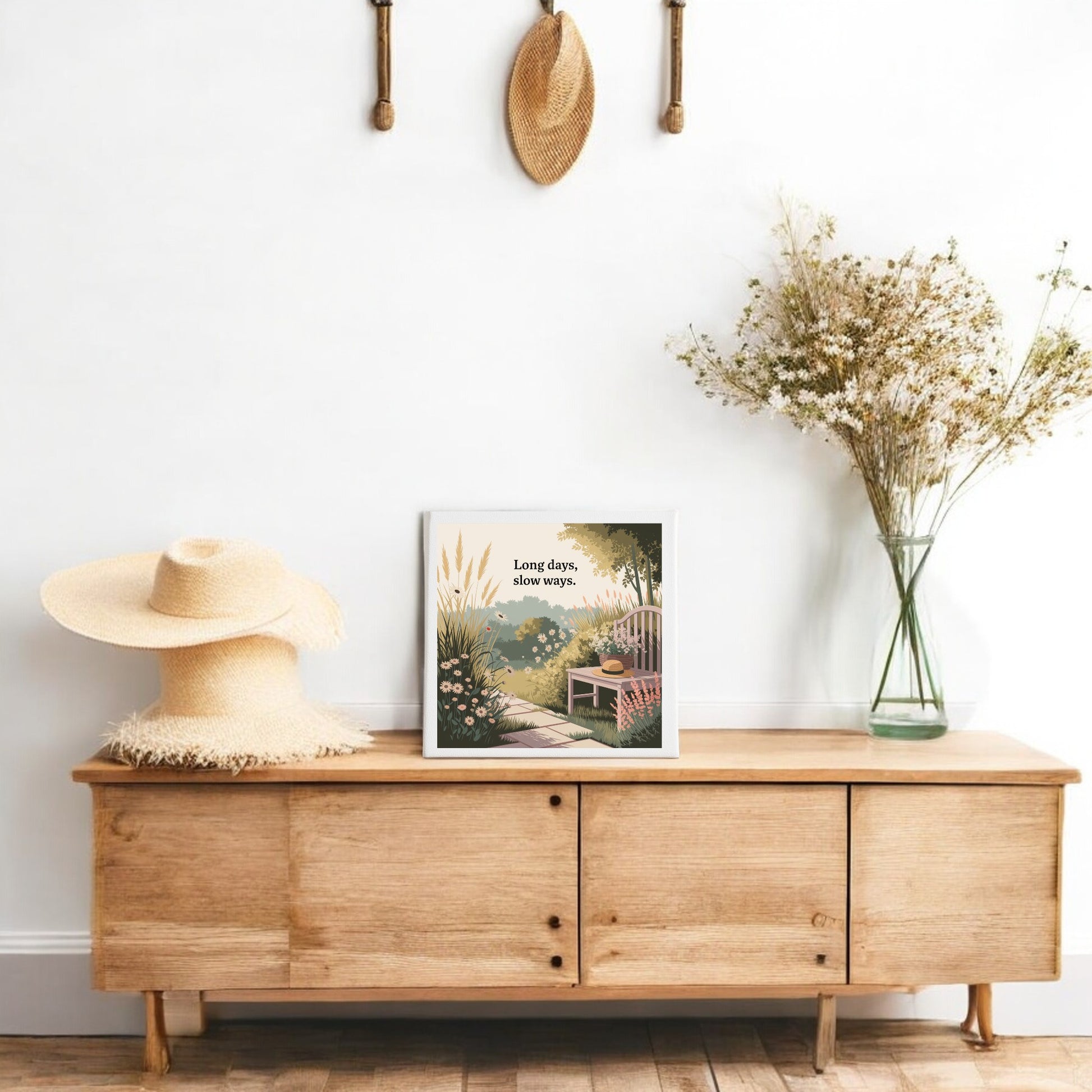 A wooden console holds a vase of dried flowers and displays "Long Days, Slow Ways Canvas – Peaceful Summer Garden Art" by Mirabilia Boutique, with straw hats above—creating calming decor for a cozy, minimalist space inspired by slow living.