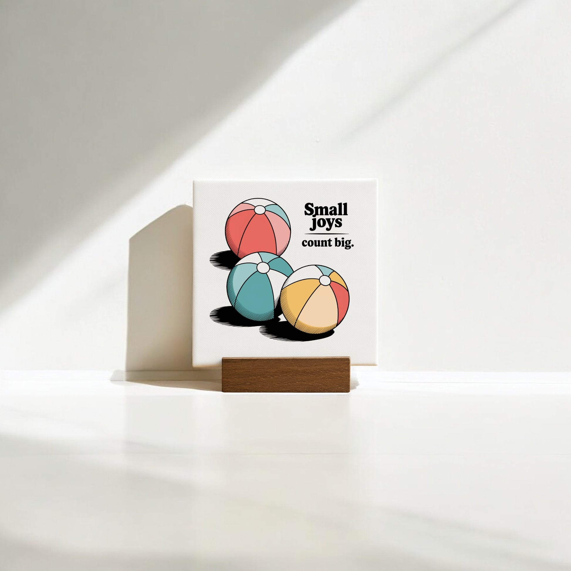 The Small Joys Count Big Mini Canvas – 4x4 Summer Desk Sign by Mirabilia Boutique displays three colorful beach balls and the phrase "Small joys count big," propped on a wooden stand, making playful summer decor for your home or desk.