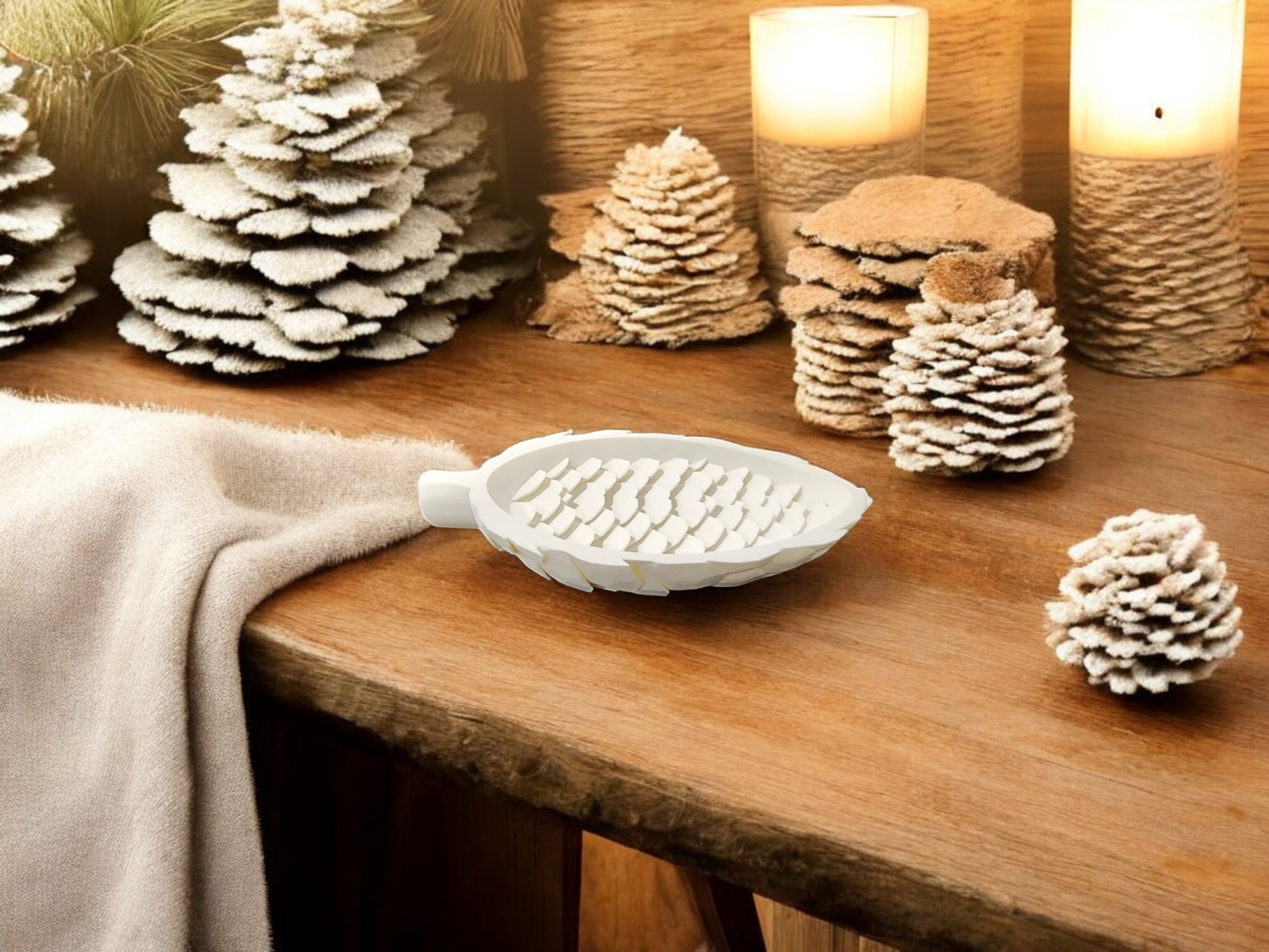 A cozy wooden table displays the Mirabilia Boutique Winter White Pinecone Tray for Woodland Winter Decor, paired with a beige blanket, lit candles, and a white leaf soap dish for a warm, rustic winter ambiance.