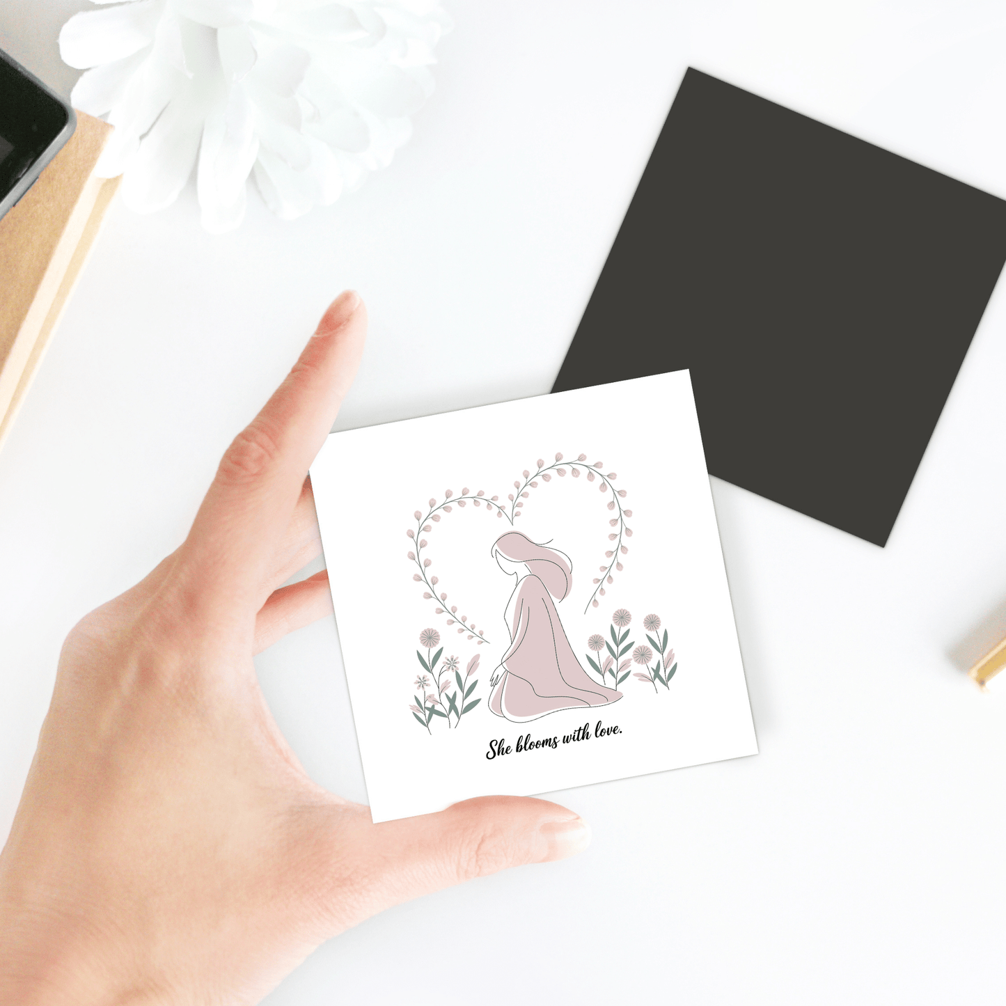 A hand holds the Mirabilia Boutique She Blooms with Love Magnet – Gentle Floral Mom Gift, adorned with a woman, flowers, and a heart-shaped vine—a thoughtful décor piece for mothers. Other items are blurred in the background.
