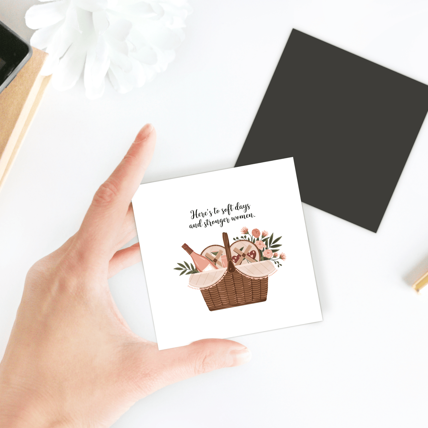 A hand holds the "Soft Days Strong Women Magnet" by Mirabilia Boutique, featuring a basket of bread and flowers with the text: “Here’s to soft days and strong women”—an empowering gift for women. A white flower accents the scene.