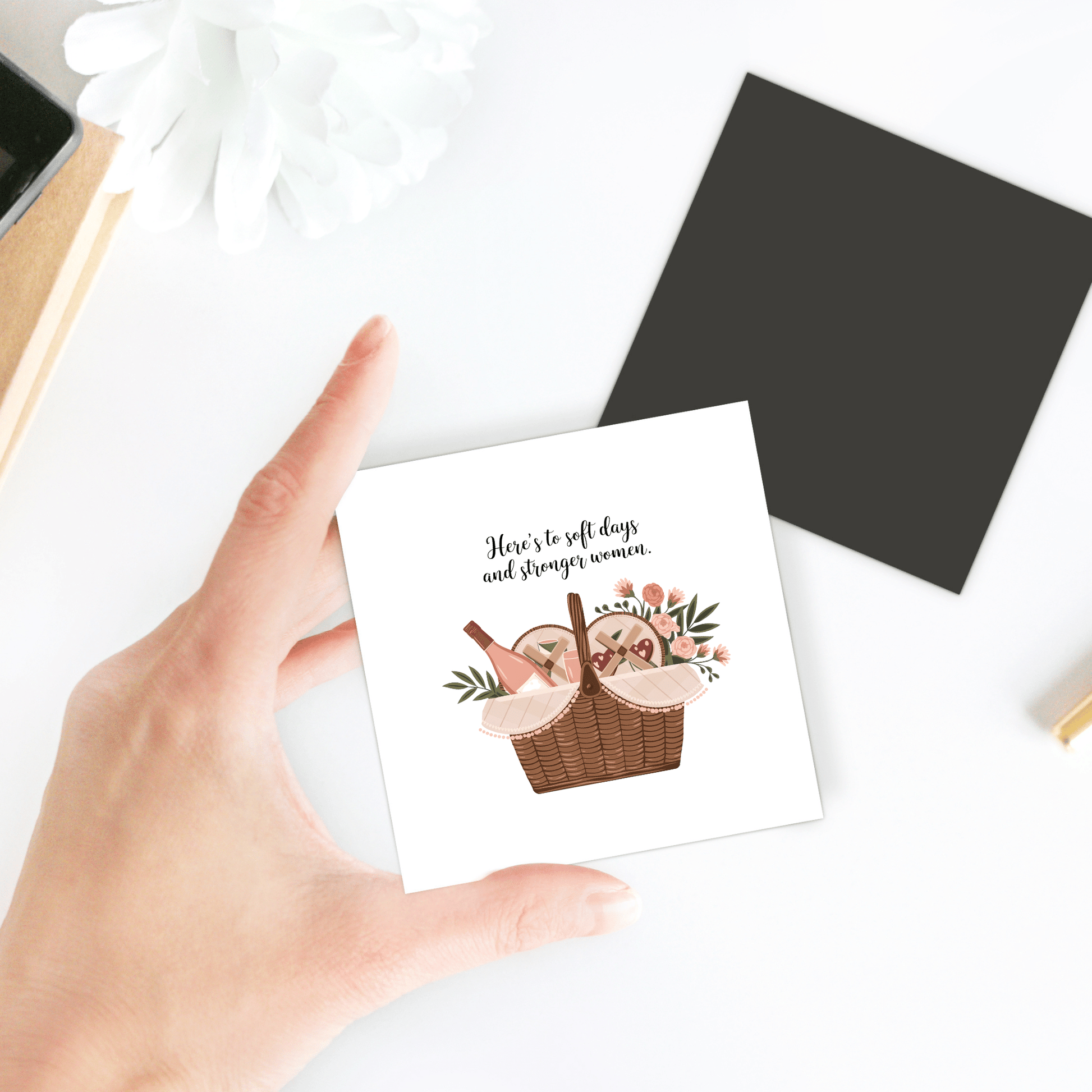 A hand holds the "Soft Days Strong Women Magnet" by Mirabilia Boutique, featuring a basket of bread and flowers with the text: “Here’s to soft days and strong women”—an empowering gift for women. A white flower accents the scene.